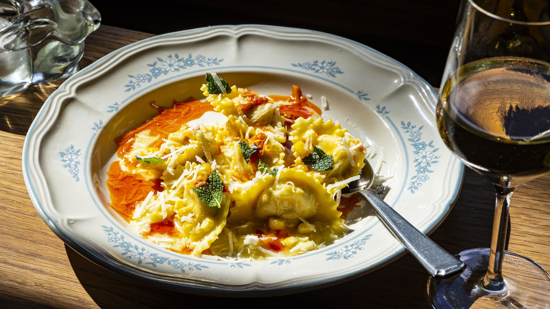 A plate of ravioli with orange sauce, cheese shavings, and mint on a blue floral-rimmed dish. A wooden table, with a glass pitcher on the left and a wine glass on the right.