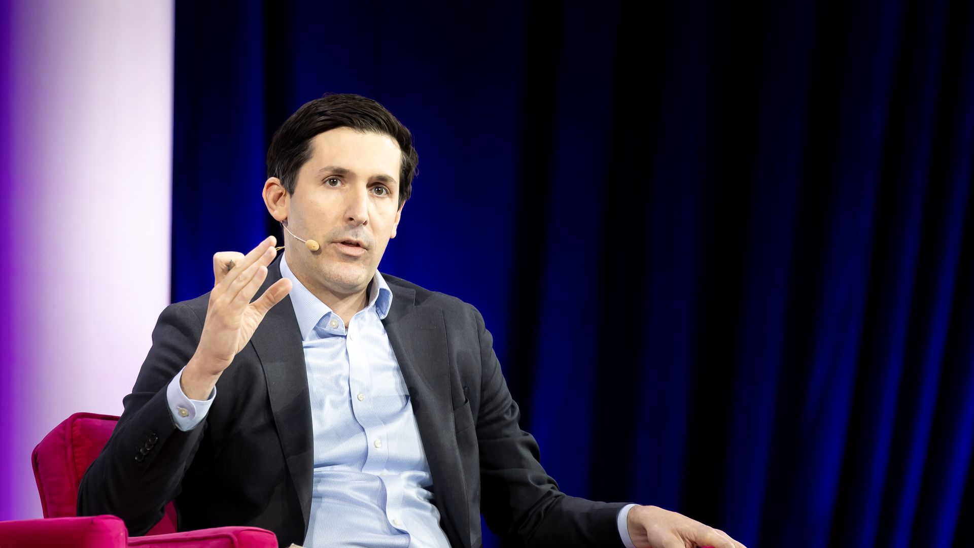 Man in a black suit and light blue shirt speaking with a headset microphone, seated on a bright pink chair against a dark blue curtain background.