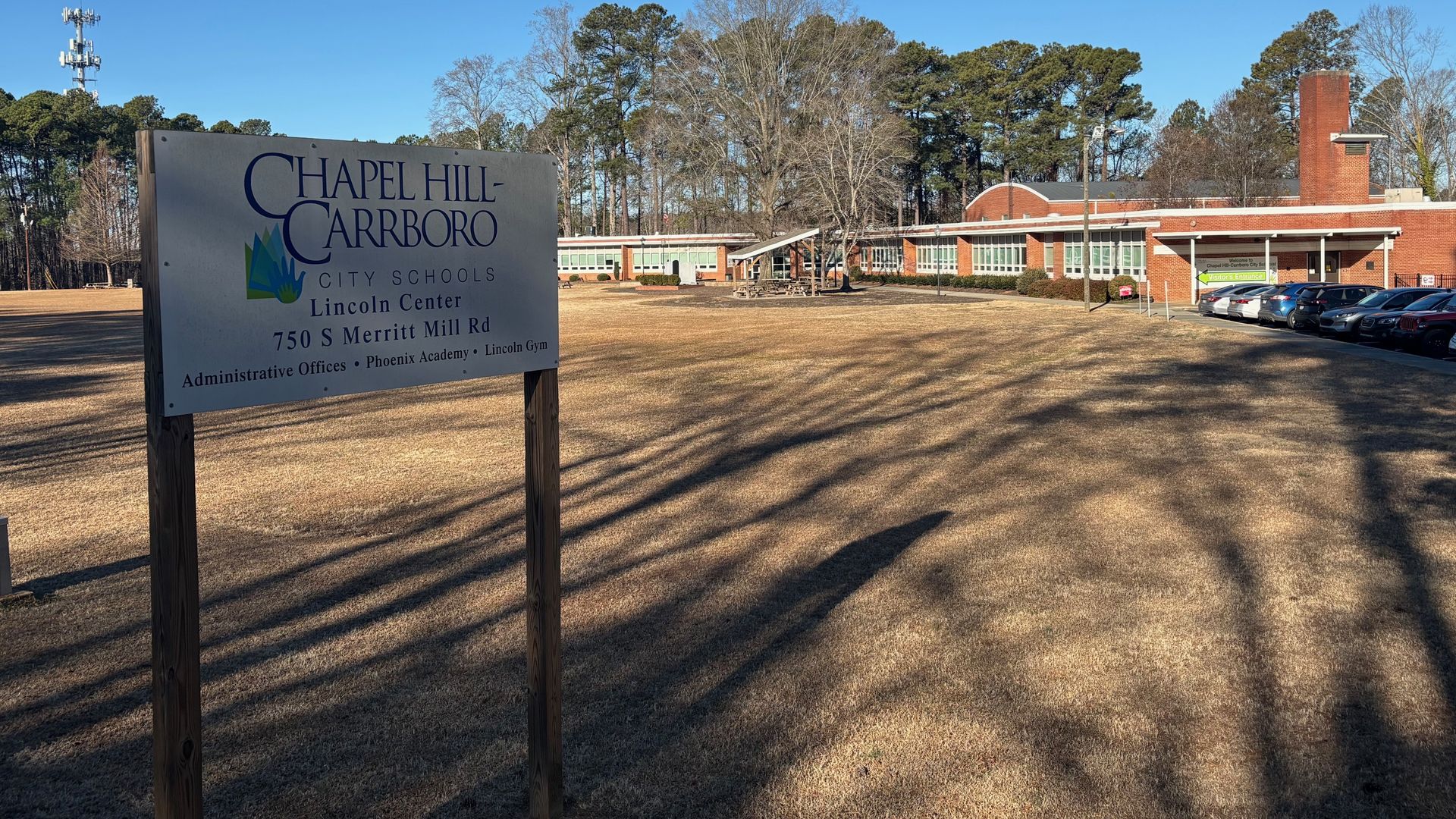 Sign for Chapel Hill-Carrboro City Schools Lincoln Center at 750 S Merritt Mill Rd in front of a brick school building with a parking lot, leafless trees, and a clear blue sky.