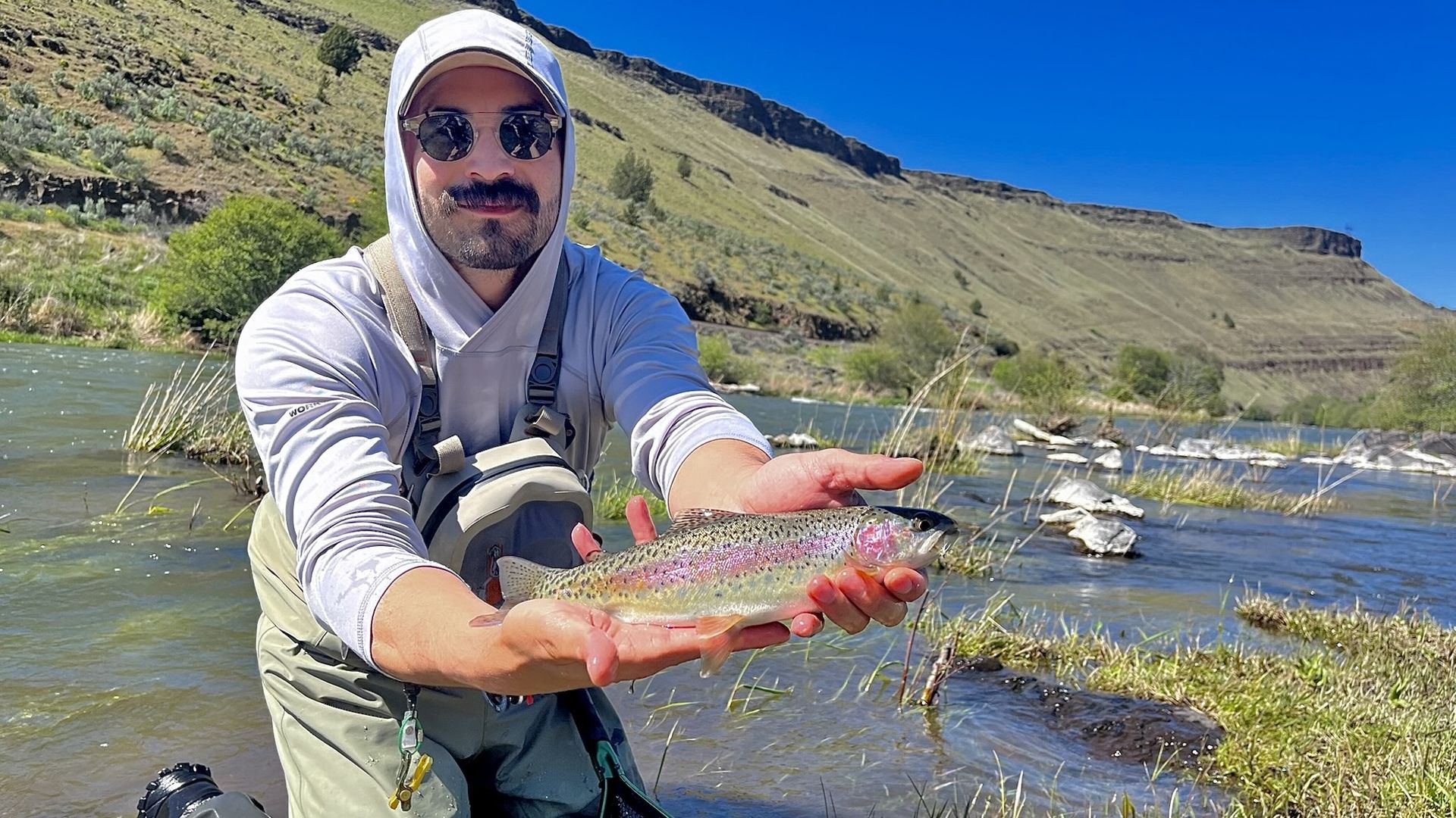 A man in fishing gear kneels by a river holding a small rainbow trout. Behind him are grassy hills, rocks, and a bright blue sky. The scene captures a peaceful, sunny day of fishing.