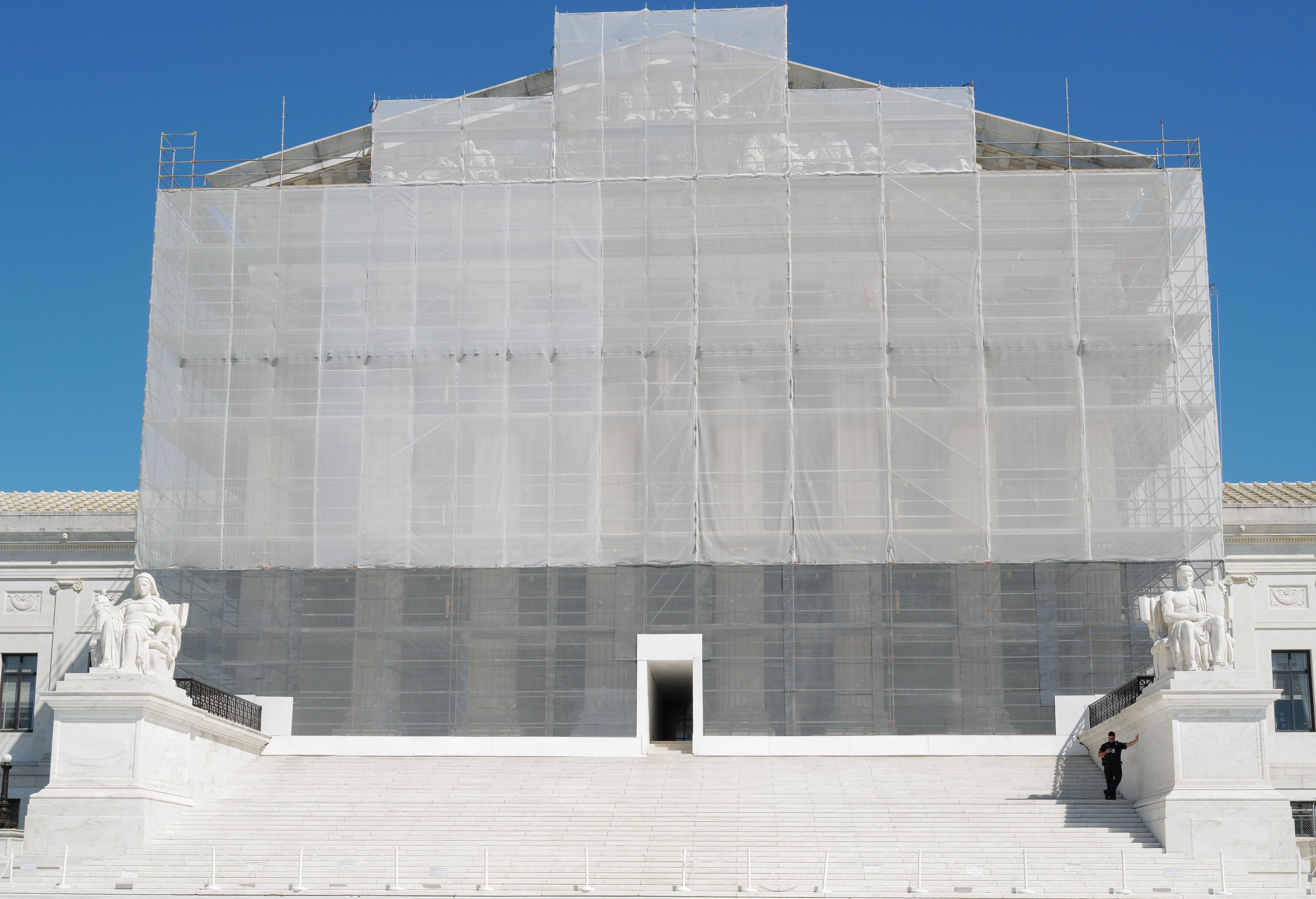White marble steps leading to a large building covered in scaffolding and white netting, with two large seated statues on either side under a clear blue sky.