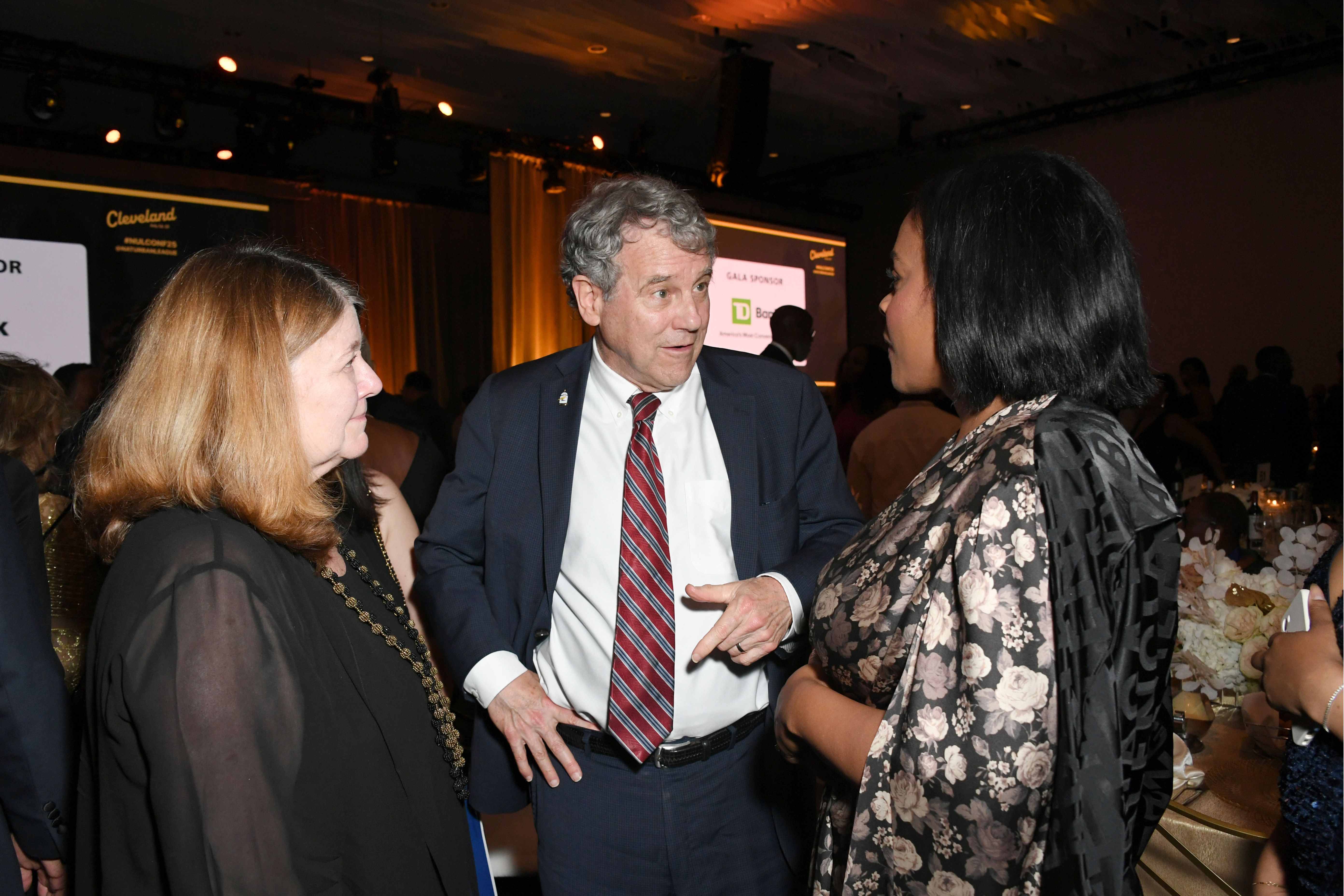 Three people engaged in conversation at an indoor gala event with dim lighting and gold curtains in the background. The man wears a navy suit and red-striped tie. Two women wear patterned and dark attire.