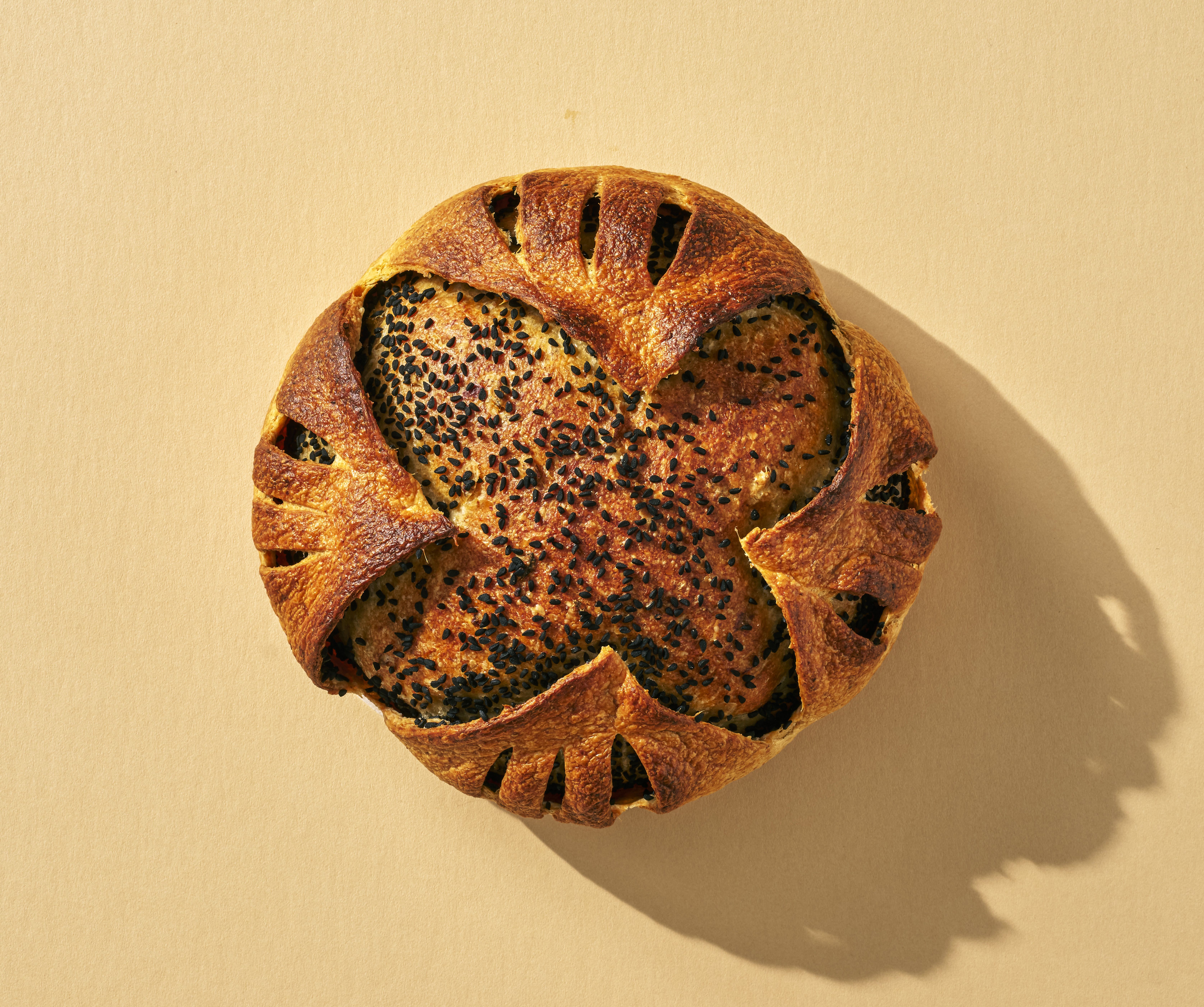 Round golden-brown bread topped with black sesame seeds and decorated with folded crust edges, casting a shadow on a beige background.