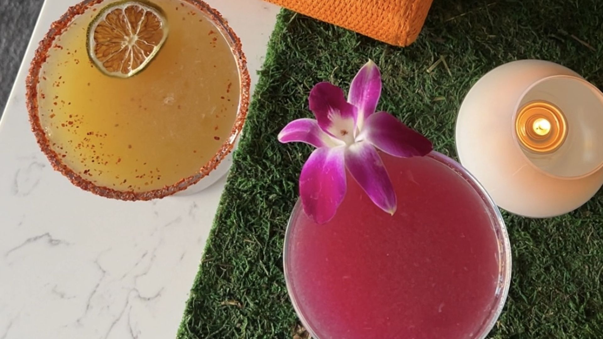 Two drinks, one yellow one pink-purple, sit on a table near a candle. 