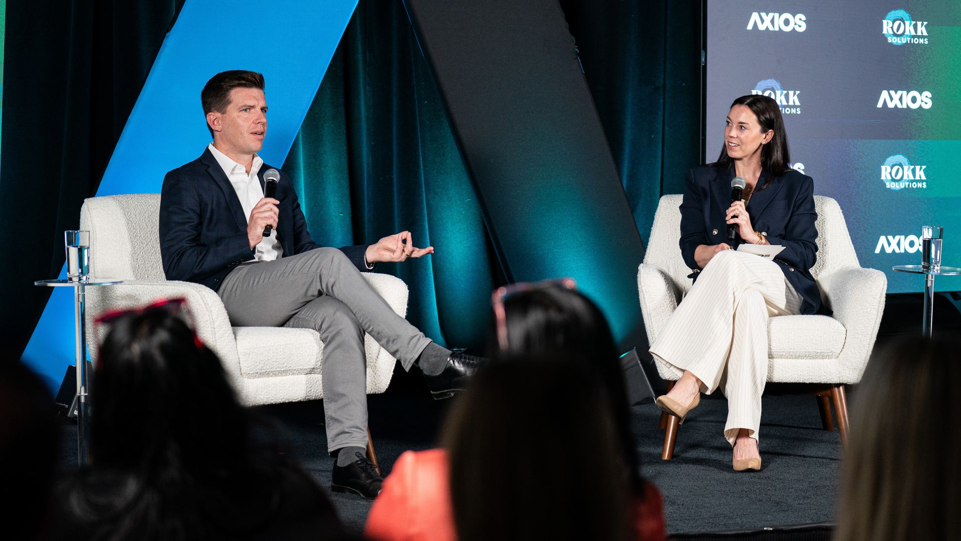Two panelists sit on stage in armchairs with microphones. Left: man in navy blazer; right: woman in dark blazer. Blue/teal backdrop featuring AXIOS logos.