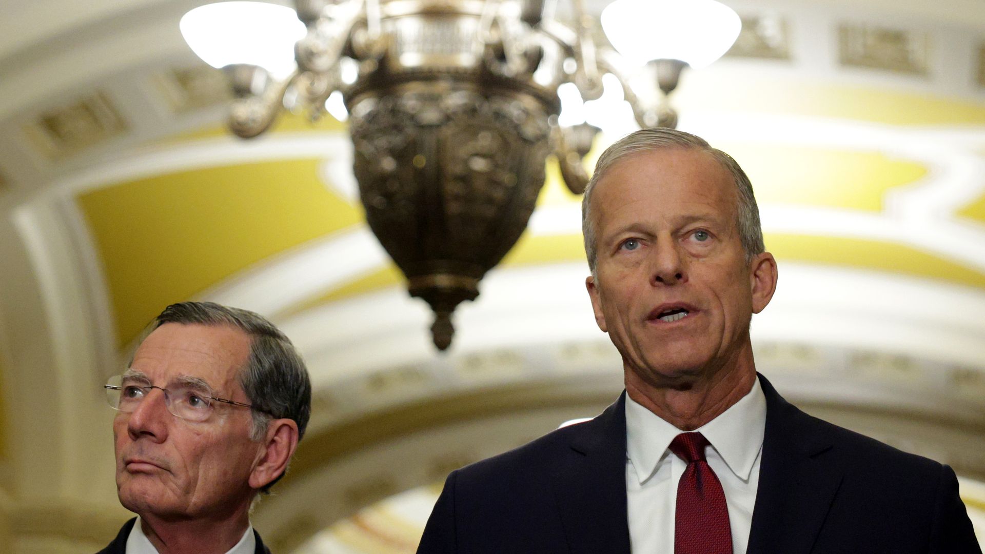 Two men in suits, one with glasses, standing under a large ornate chandelier in a room with yellow and white arched ceiling details, both looking serious during a speech or announcement.