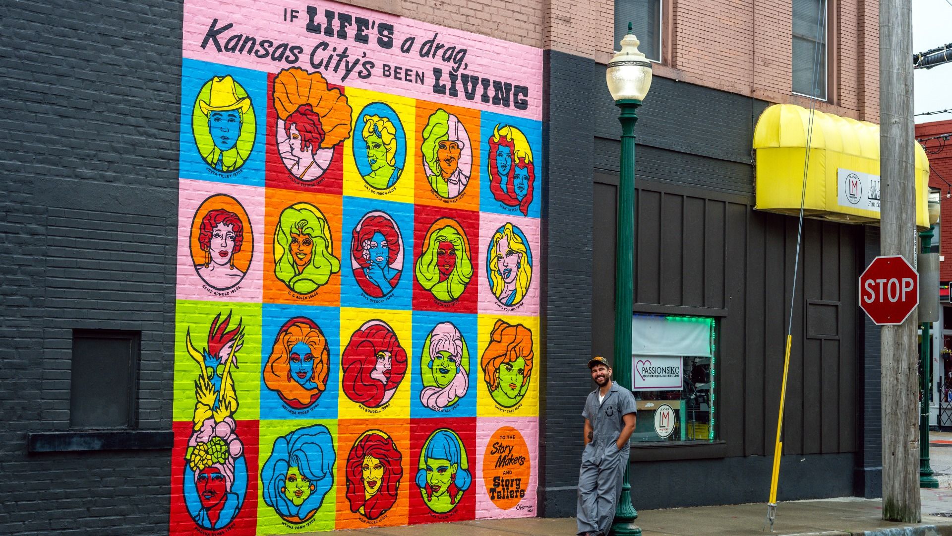 Colorful mural on a brick wall features a grid of pop-art portraits in bright squares with bold text along the top. A man in gray overalls stands by a green lamppost near a yellow awning and a STOP sign.
