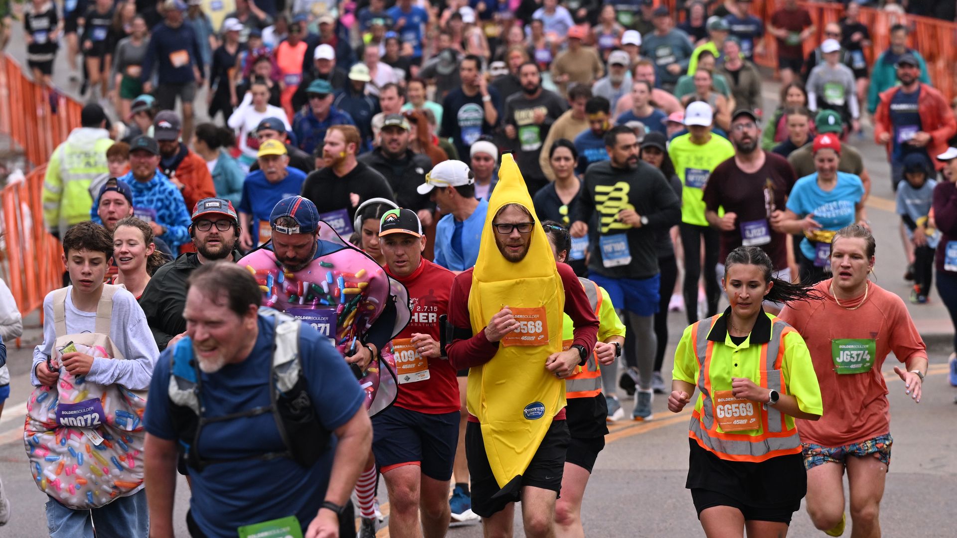 A crowded street race with hundreds of runners wearing bibs and colorful outfits; a central runner is in a bright yellow banana costume, as orange barriers line the road.