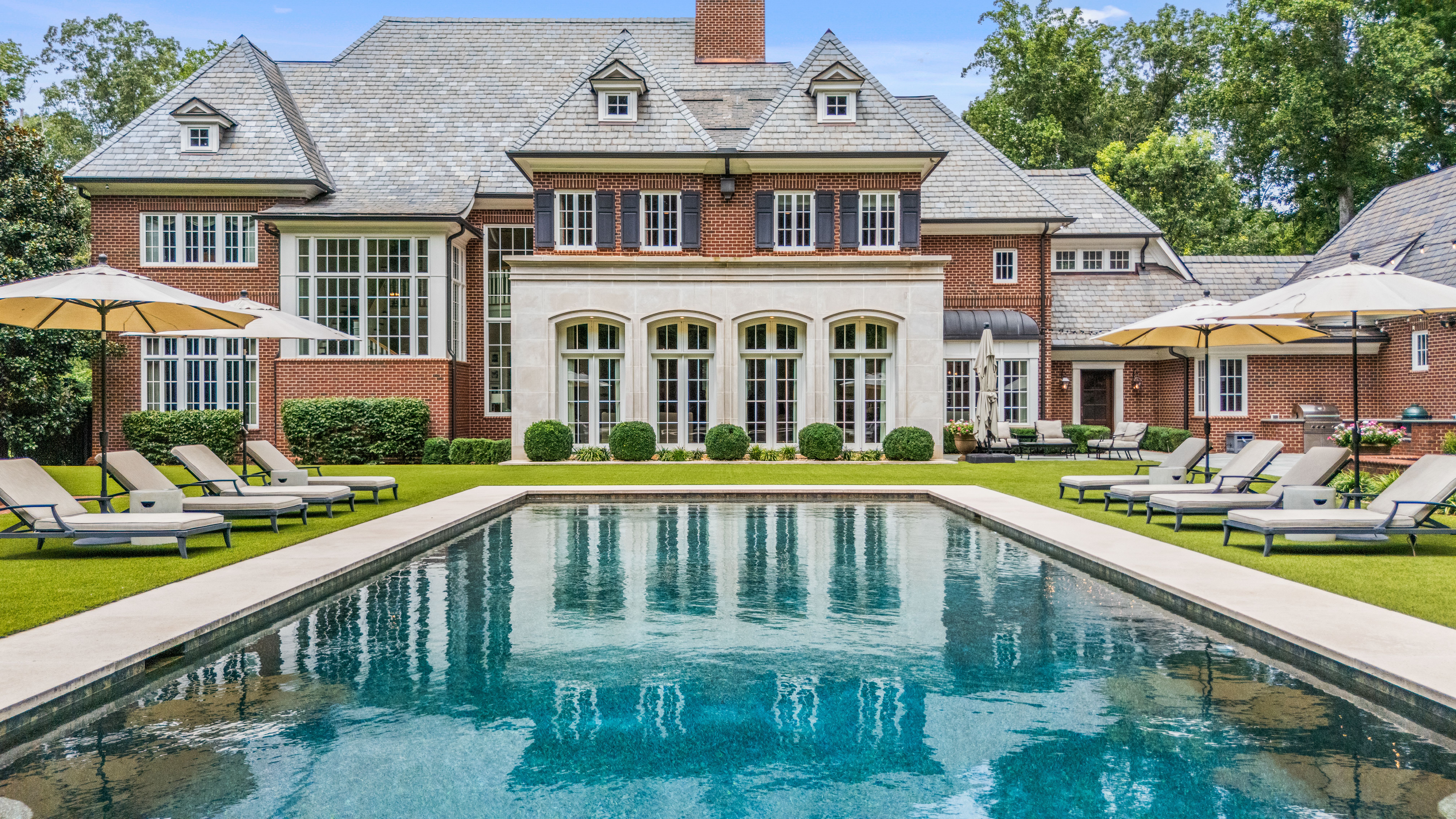 Large brick mansion with slate roof and tall windows, reflected in a rectangular swimming pool. Poolside lined with beige lounge chairs and large beige umbrellas on green lawn.