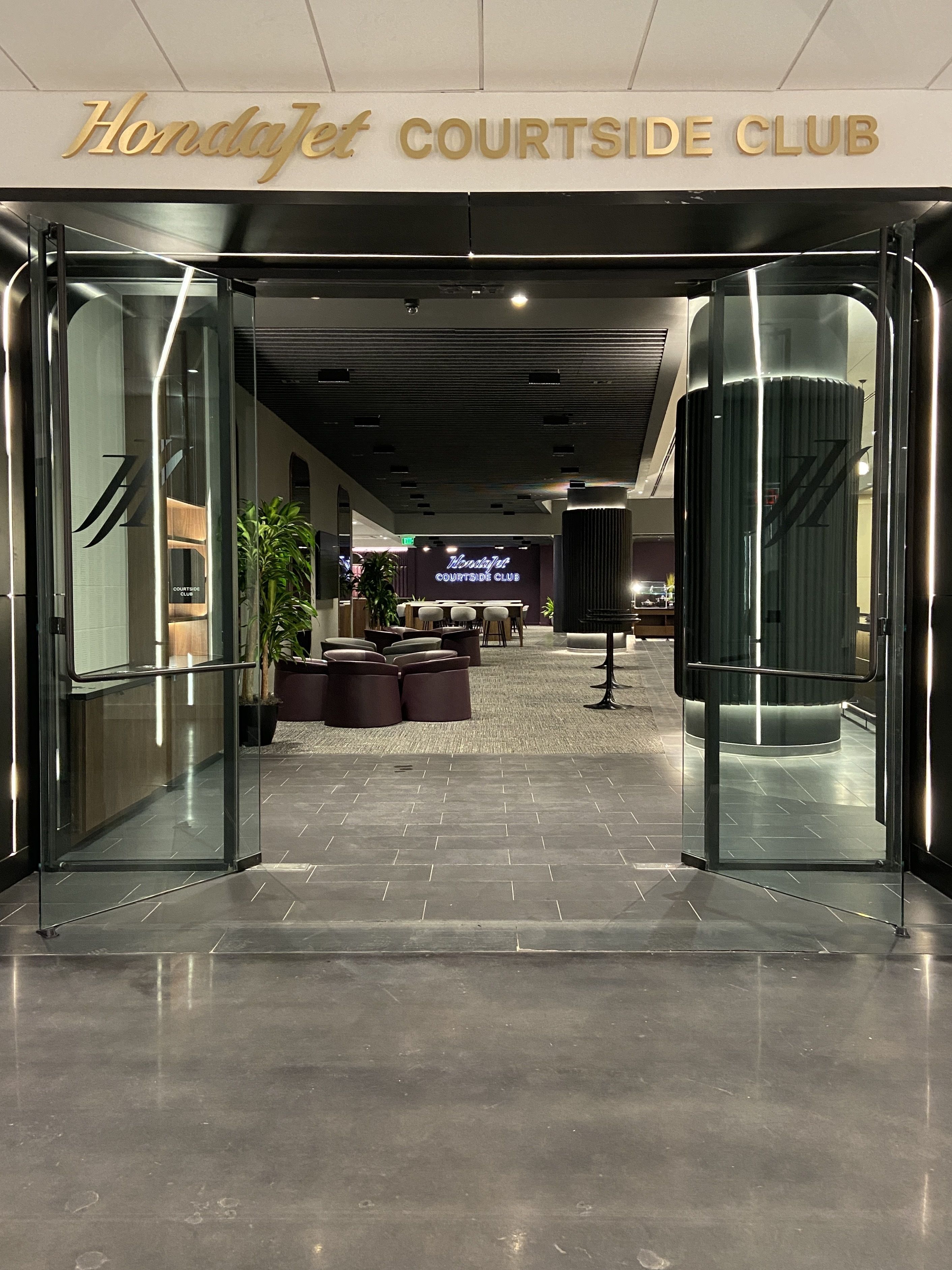 Entrance to the HondaJet Courtside Club with glass doors, polished stone floor, modern seating, plants, and a neon sign inside displaying "HondaJet Courtside Club."