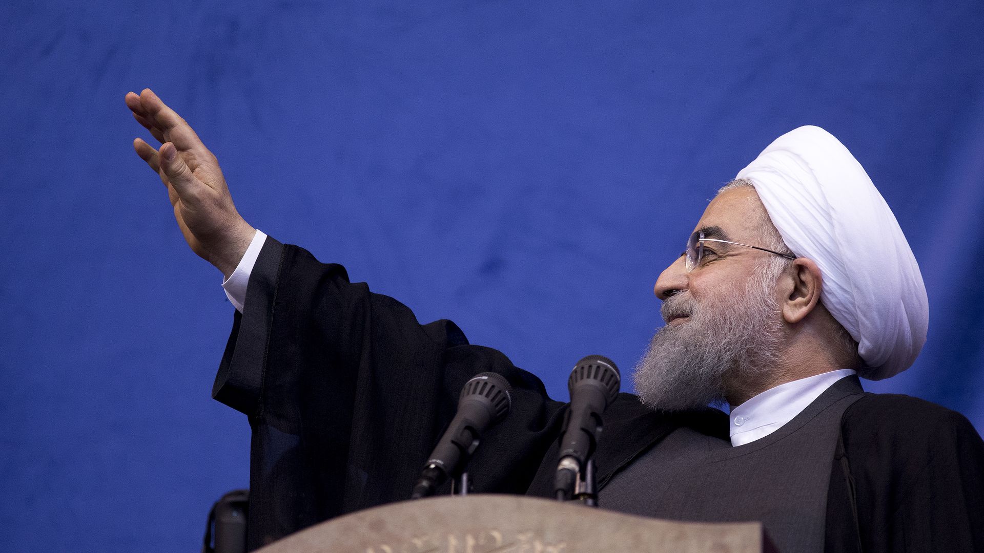 Hassan Rouhani waves his hand up before a blue background.