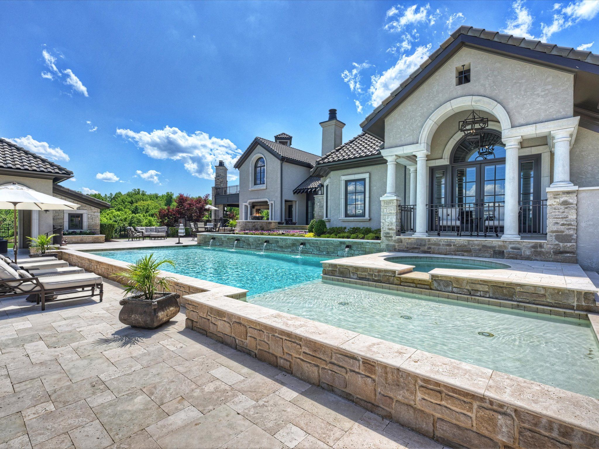 Luxury backyard with a large turquoise swimming pool, hot tub, stone patio, lounge chairs, and a cream-colored house with columns under a bright blue sky with clouds.
