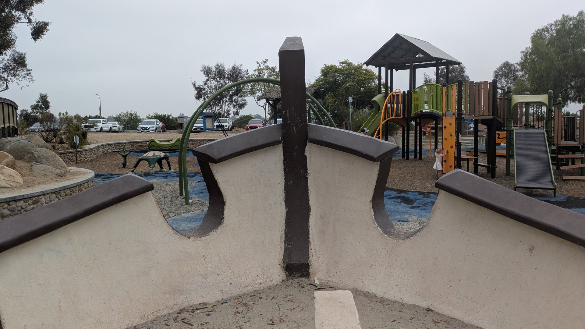 Wide view of a playground: curved concrete walls surround a central dark pillar; to the right are green and orange play structures. Trees and parked cars line the background under a gray sky.