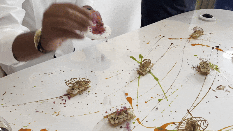 A chef tosses garnish onto a white table filled with artistic edible bites.