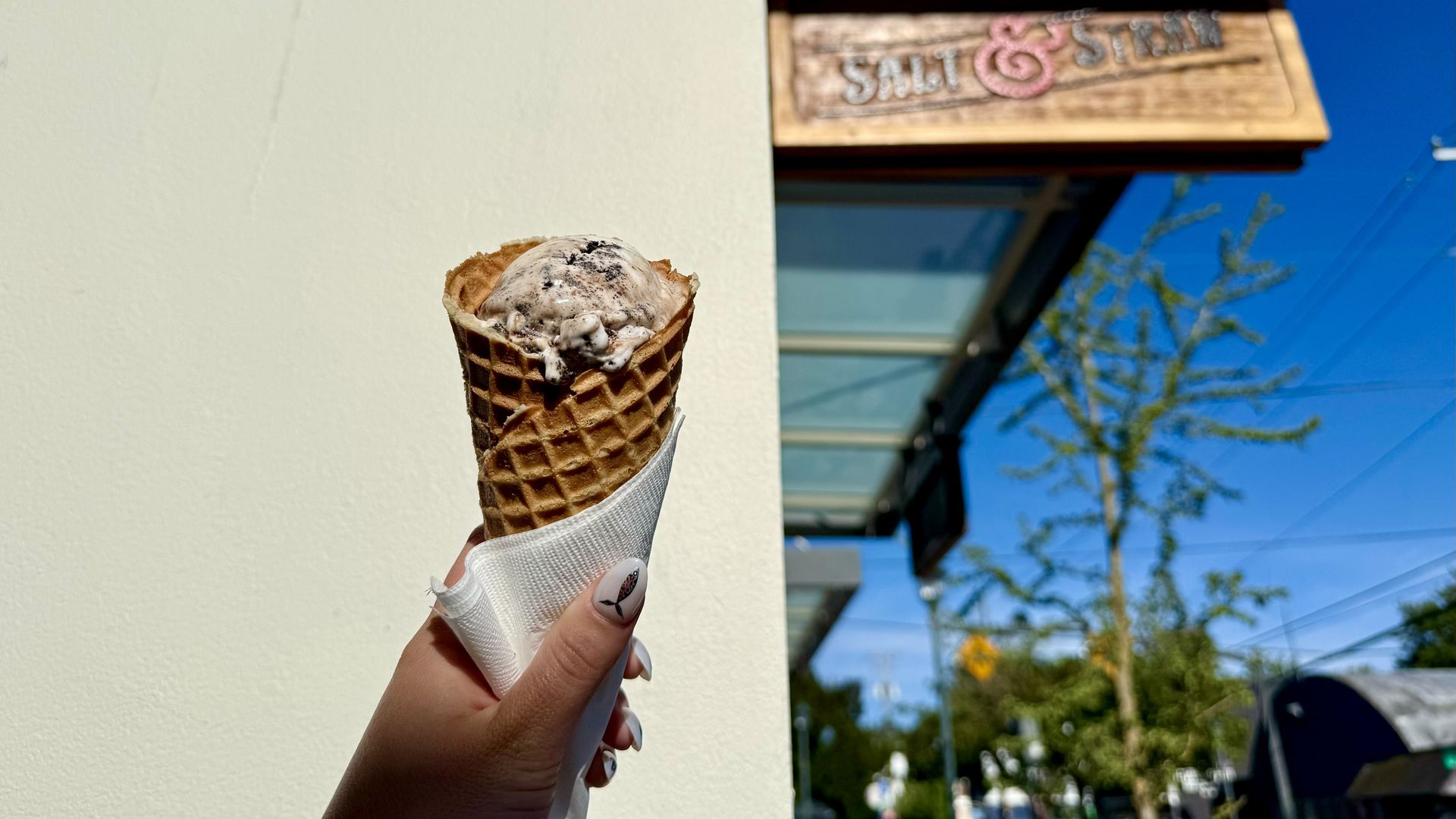 Hand holding a waffle cone with cookies and cream ice cream against a light wall, with a blurred outdoor background featuring a wooden sign that reads "Salt & Straw".