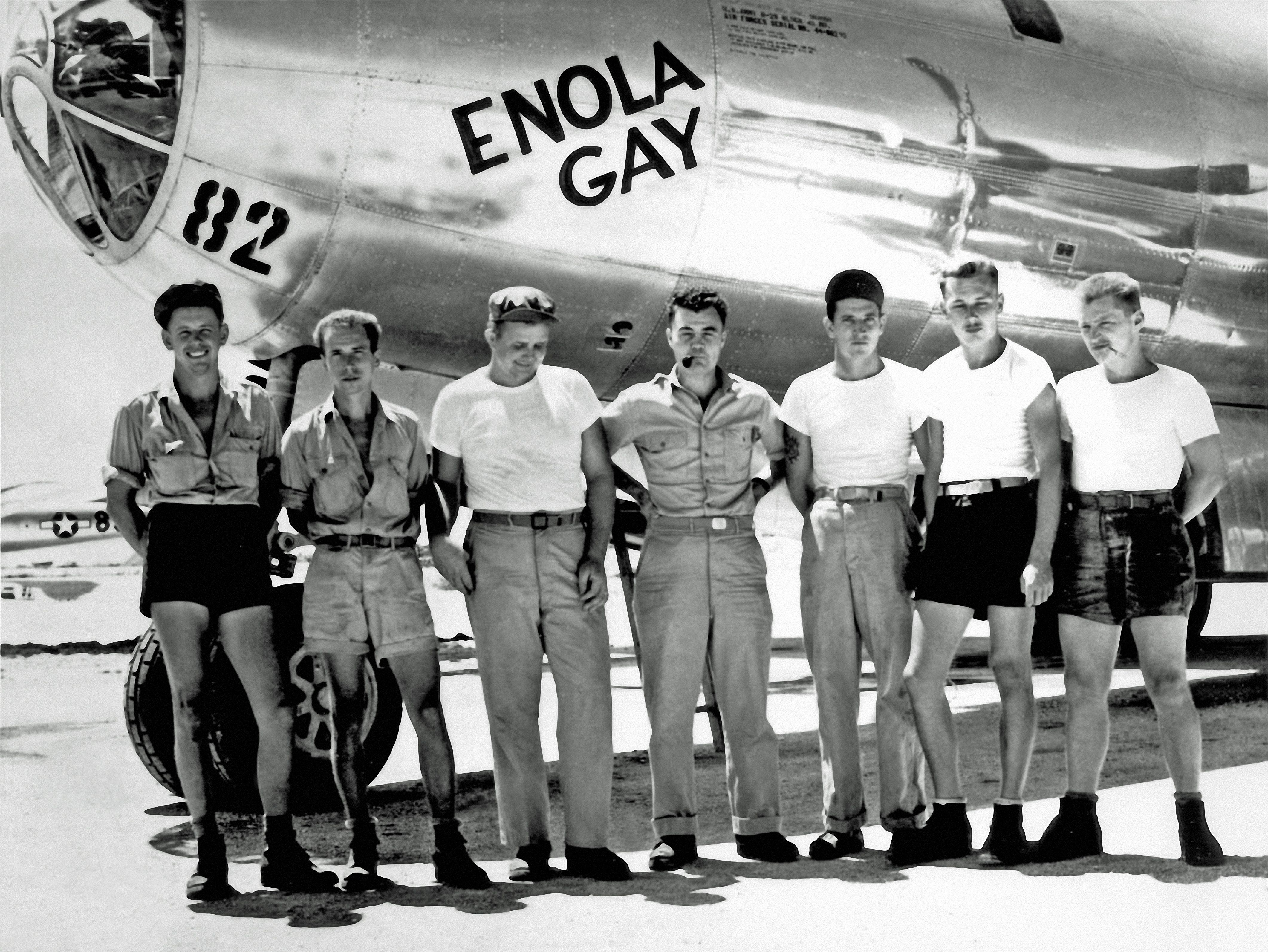 Black and white photo of seven men in casual military attire standing in front of the nose of the airplane "Enola Gay" with the number 82 visible on it.