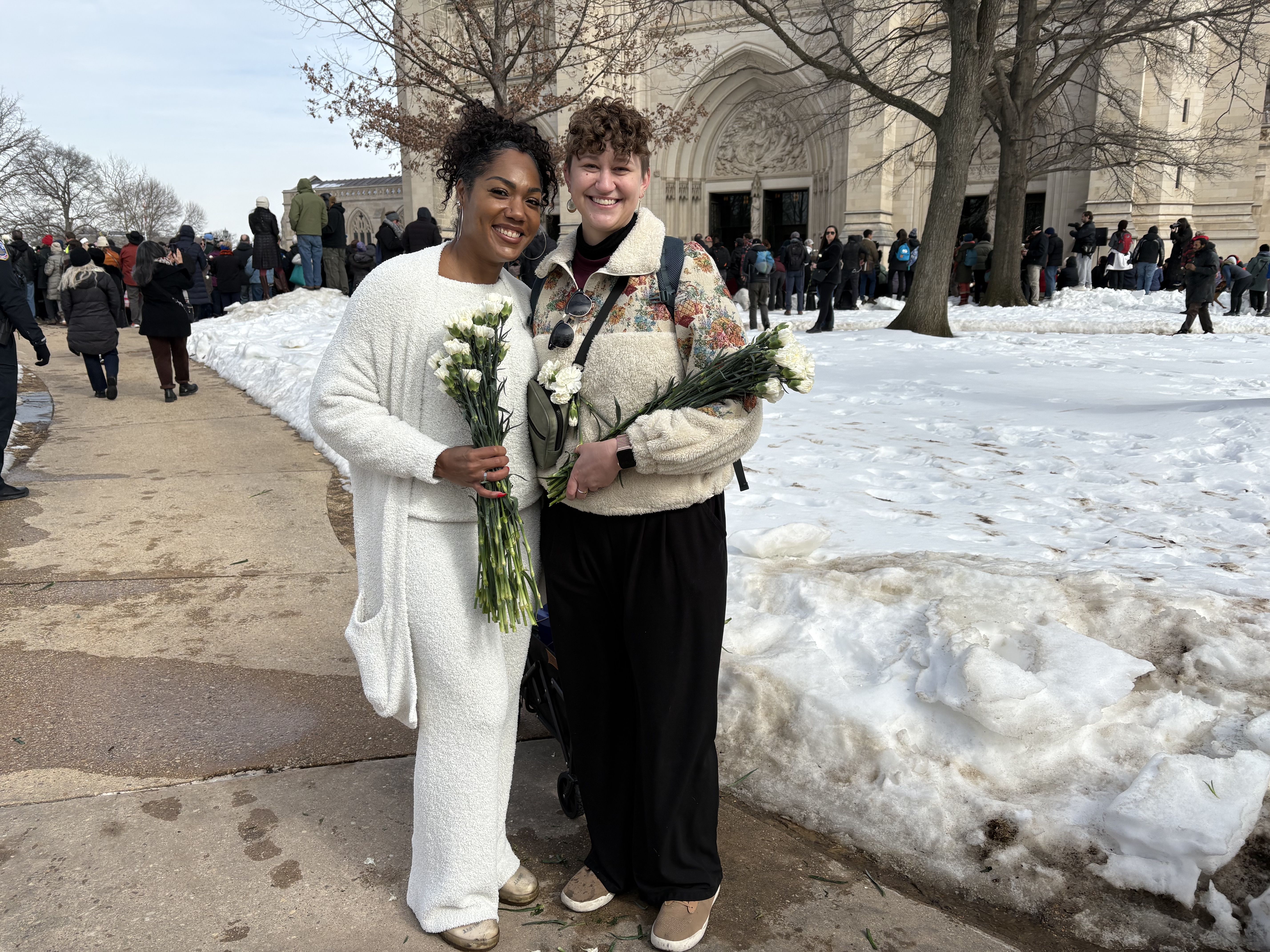 Two smiling women in winter clothes holding white flowers stand on a snow-covered path outside a large stone building with a crowd gathered nearby.