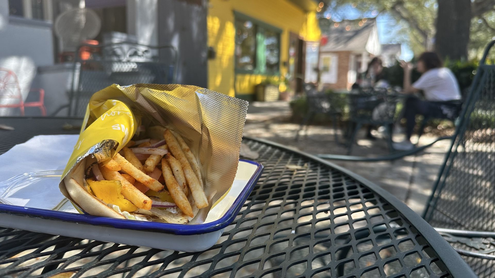 A gyro stuffed with French fries sits on a small white metal tray with blue trim. The plate is on an outdoor table, and the bright yellow restaurant Smoke & Honey can be seen in the background.