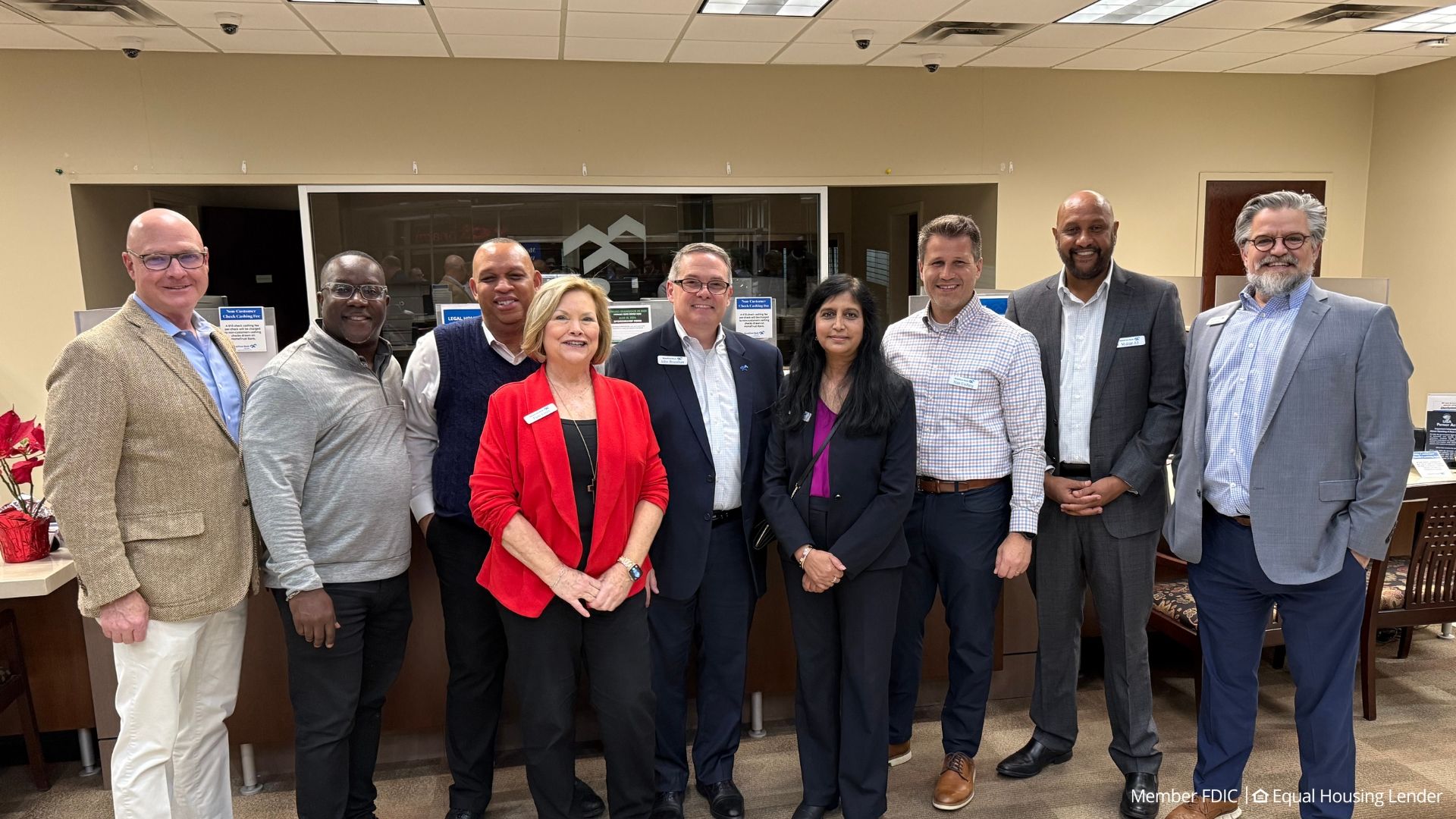 Nine professionals in business attire pose in a beige office lobby with a glass wall behind. A woman in a red blazer stands center; others wear navy, gray, and light blue. FDIC watermark bottom right.