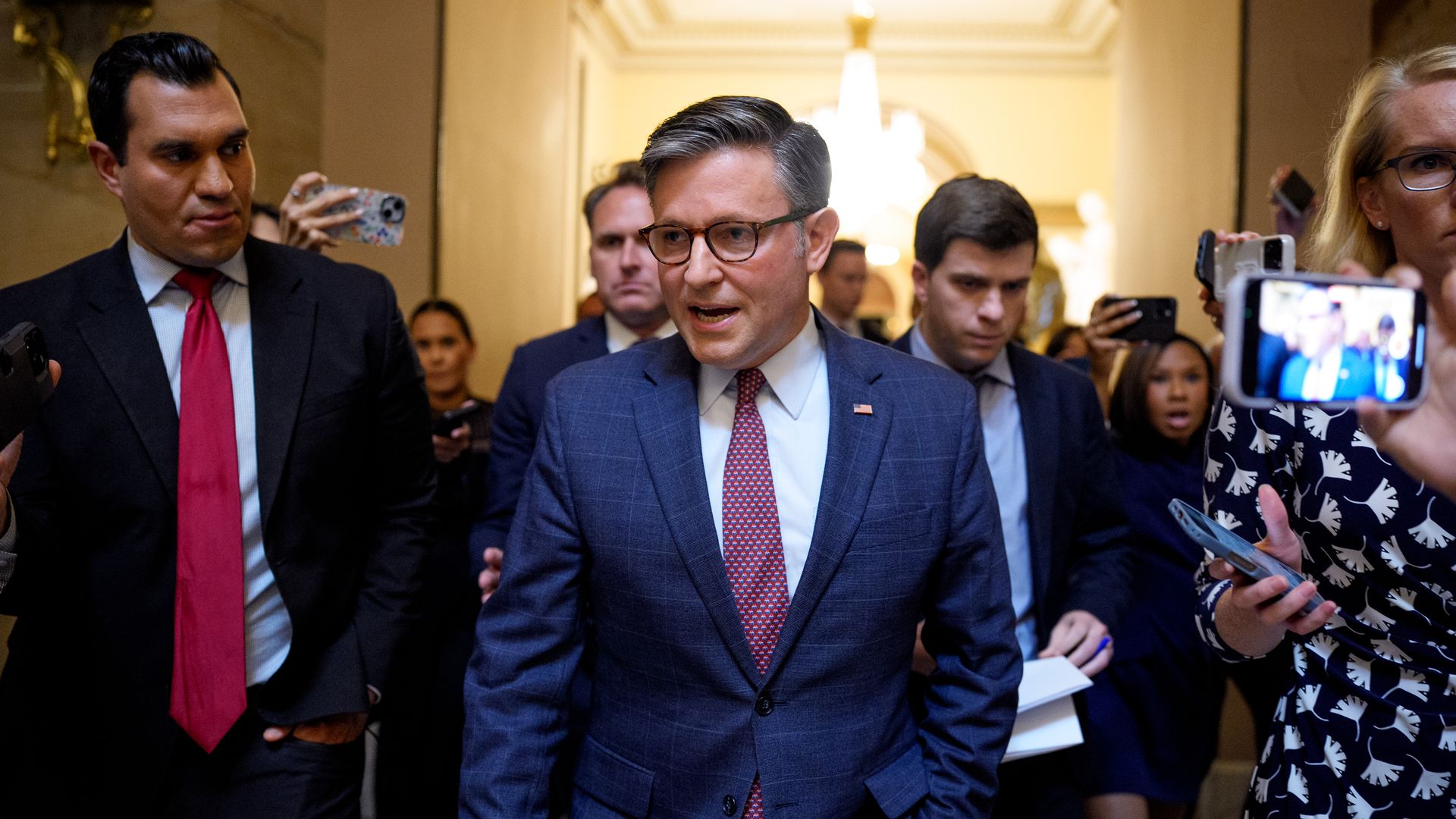 Mike Johnson talks as he walks on Capitol Hill in a blue suit while reporters film him on their phones.
