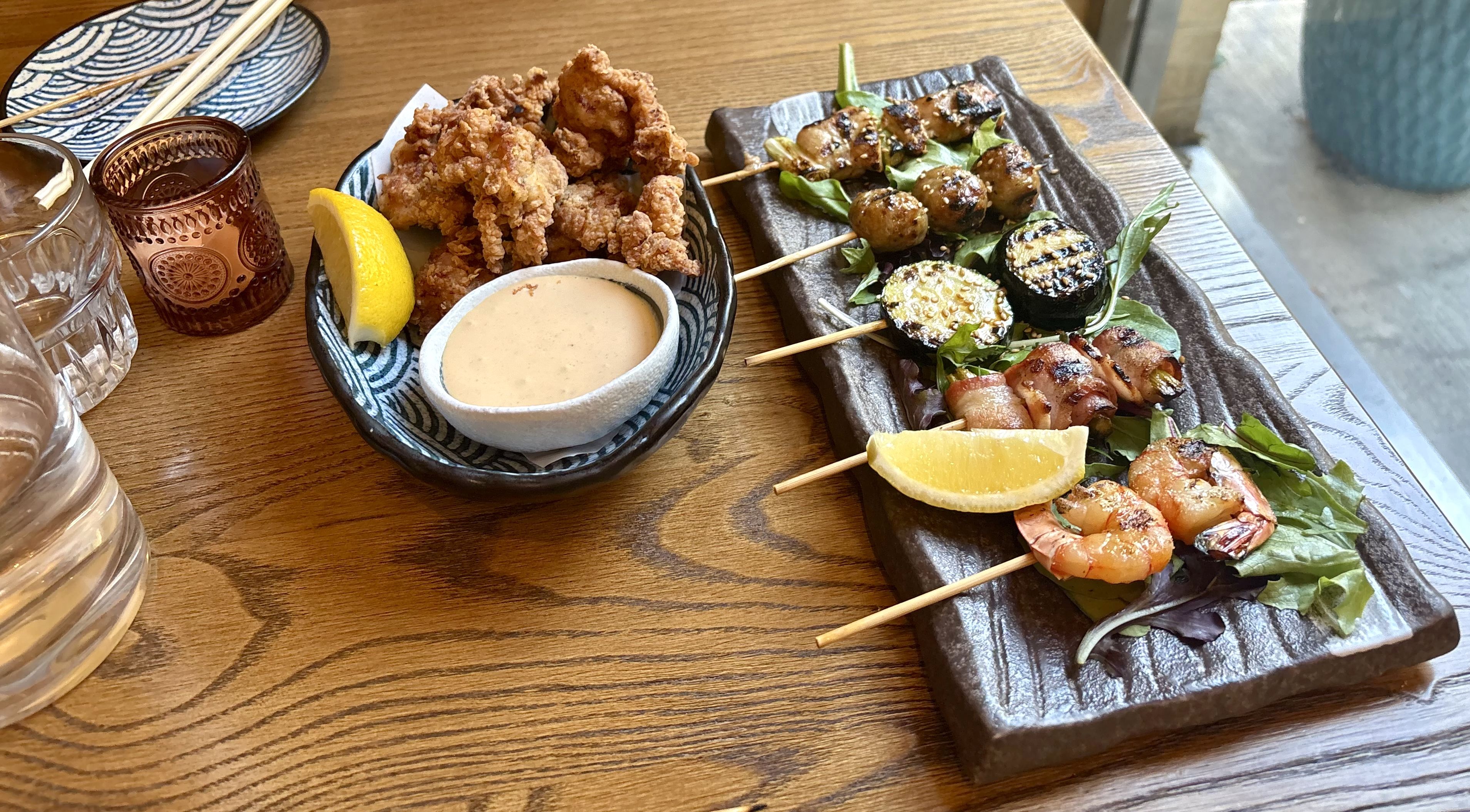 On a wooden table, a blue patterned bowl holds fried chicken bites with a lemon wedge and dipping sauce; beside it a dark slate platter features grilled skewers with meat, vegetables, greens, and lemon.