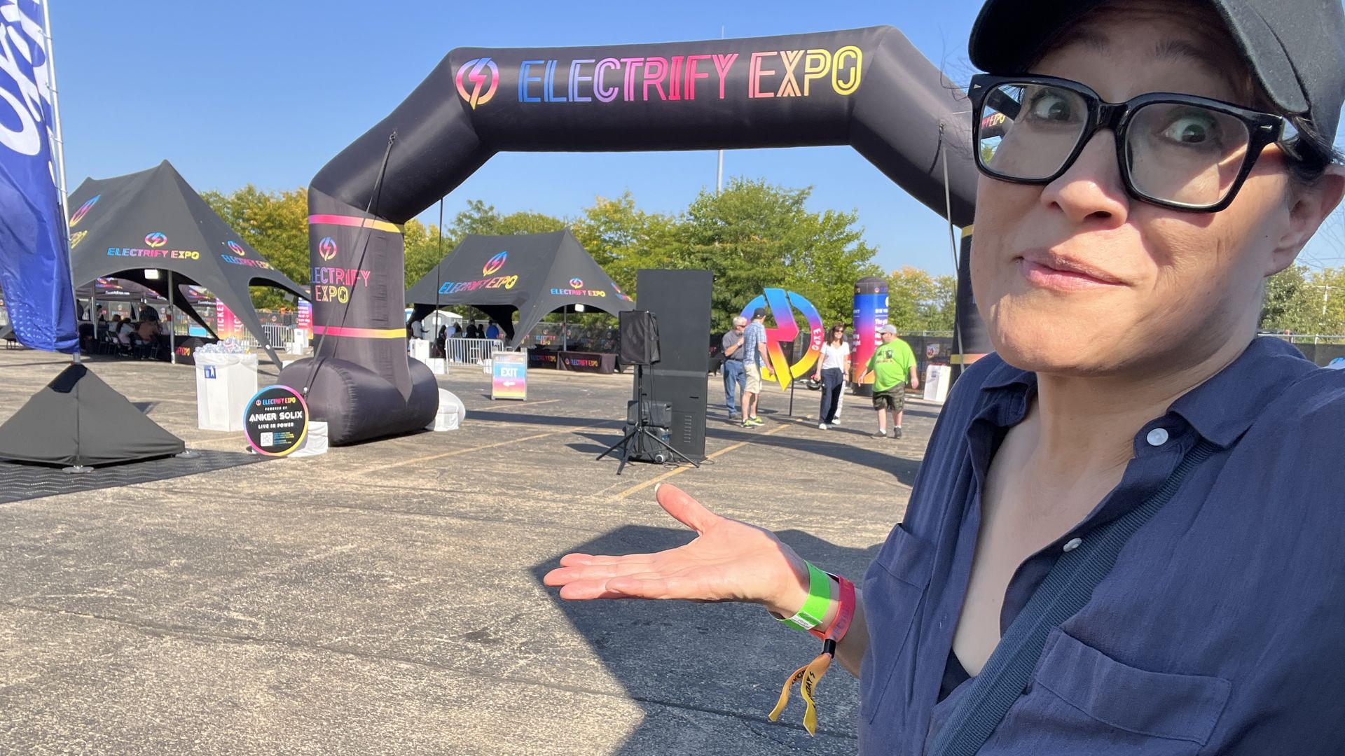 Person with glasses and a black cap shrugs in front of an entrance arch reading "ELECTRIFY EXPO" with tents and people in the background under a clear blue sky.