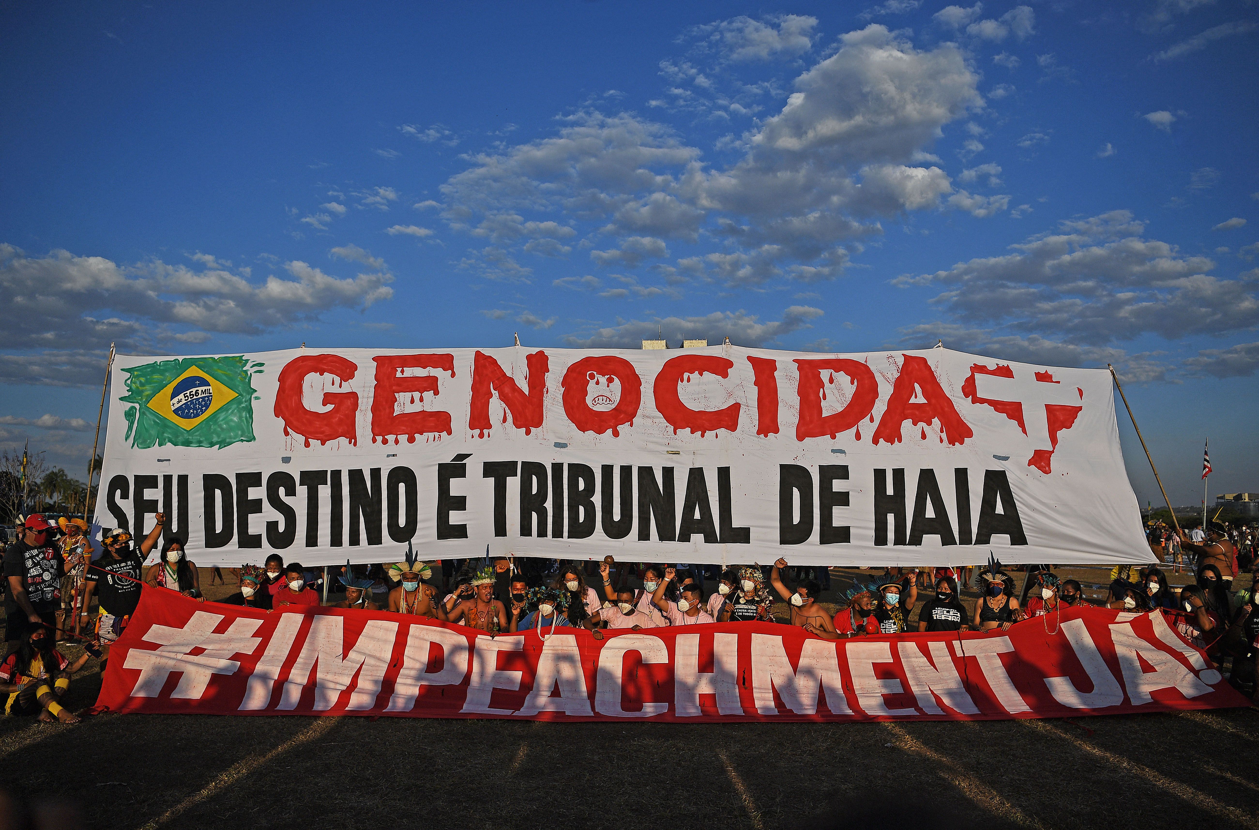 Photo of Indigenous people holding up 2 banners that say: "Genocides, your fate is the Hague court" and "Impeachment" in Portuguese