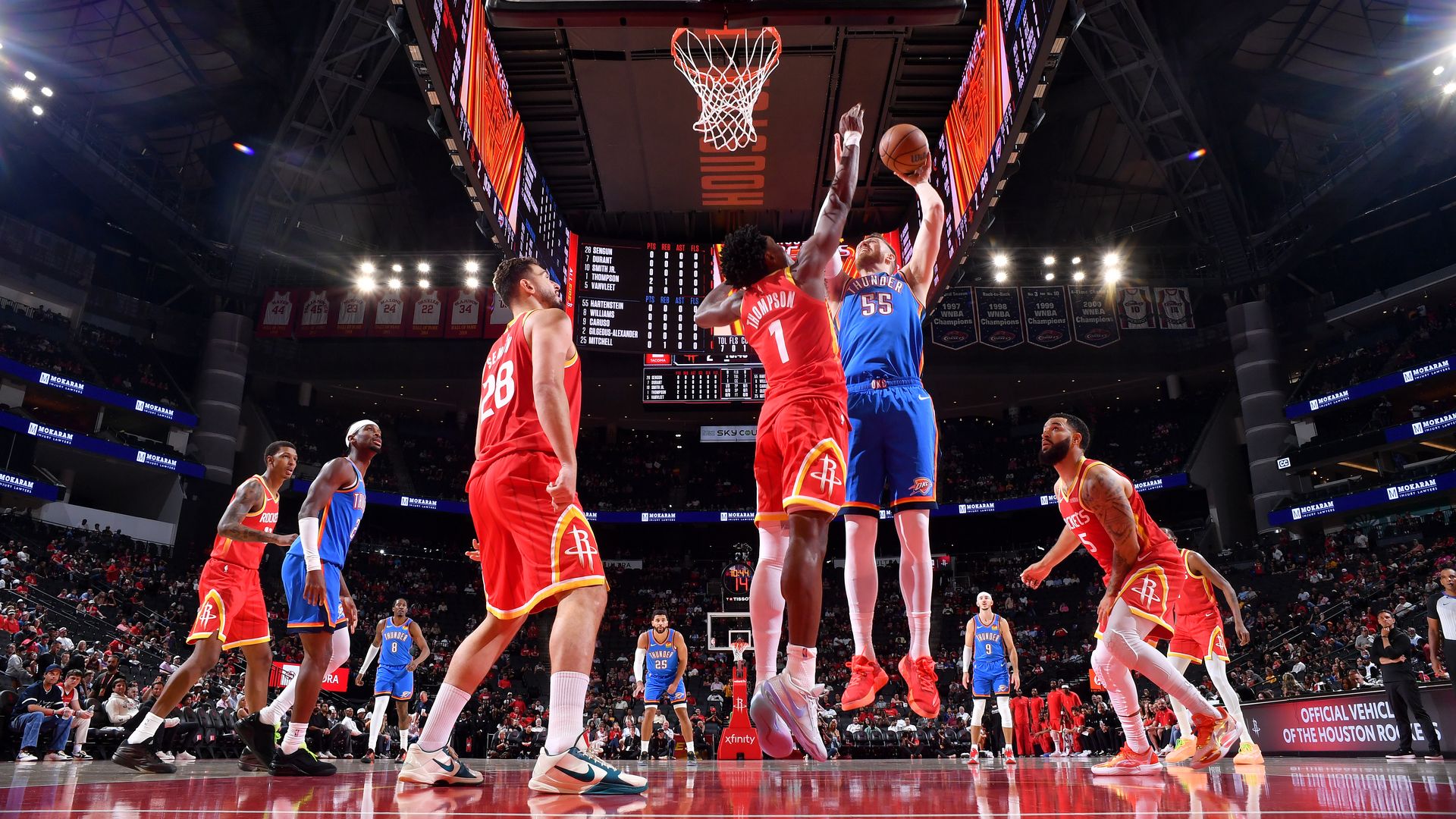 Basketball game in a packed arena: Houston Rockets in red jerseys guard an Oklahoma City Thunder player in blue as they leap for a layup near the rim; bright scoreboard and arena lights loom overhead.