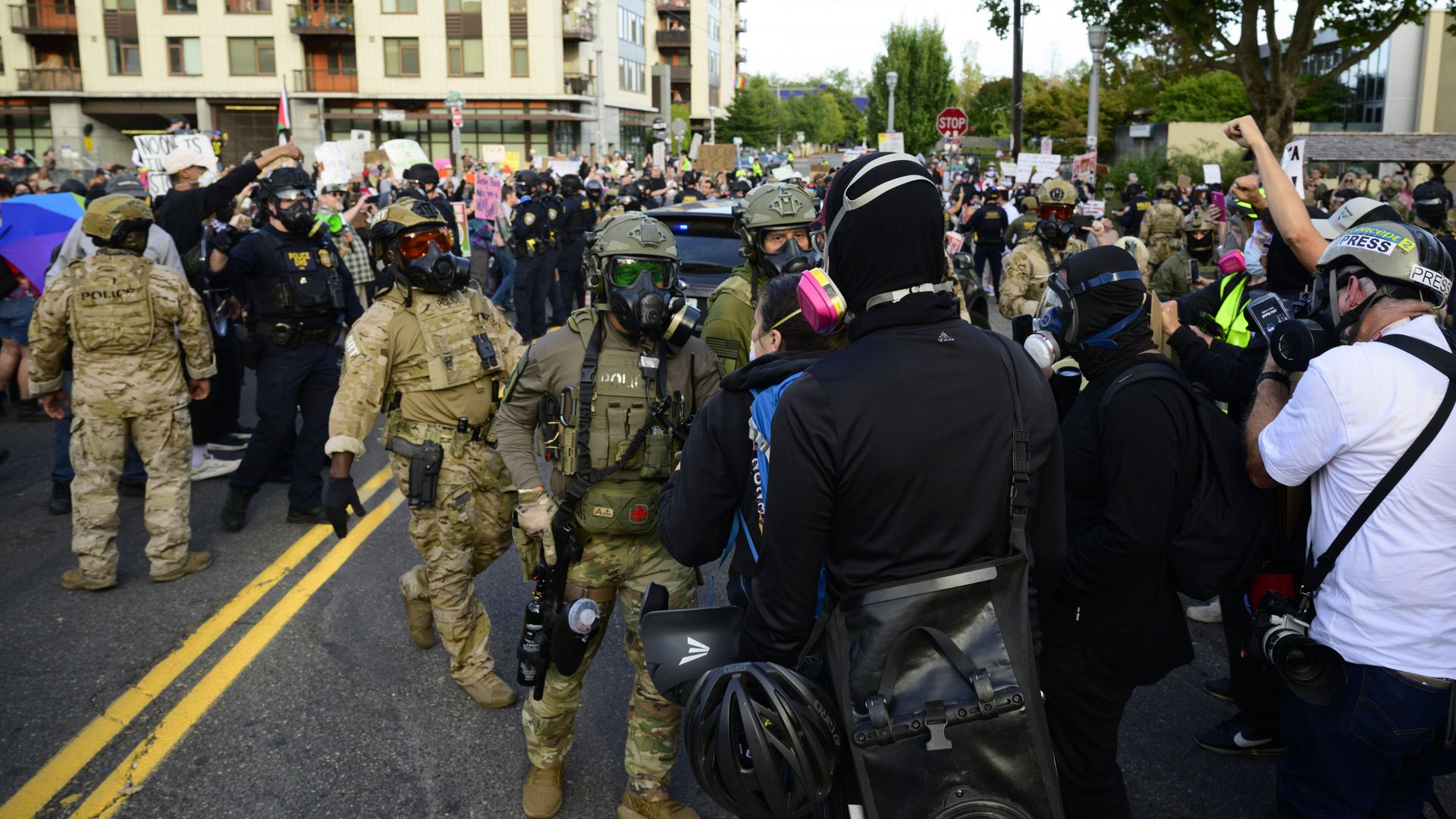 Crowd of armored police and gas-masked officers confronting protesters and press wearing gas masks and helmets on a city street during a demonstration with signs visible in background.