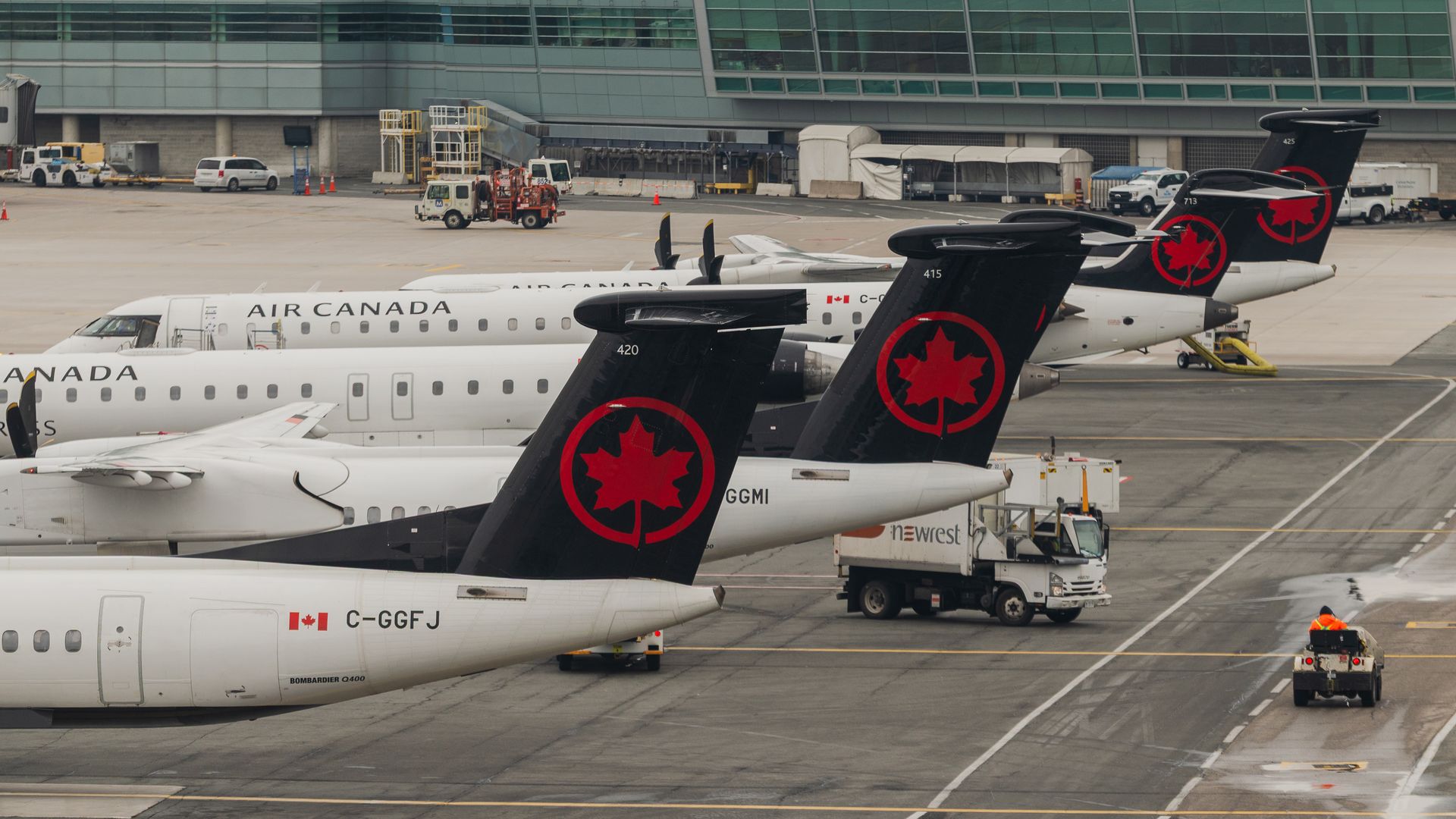 Row of Air Canada aircraft parked at the gate, black tails with red maple leaf logos, white fuselages, and ground support vehicles with a modern terminal in the background.