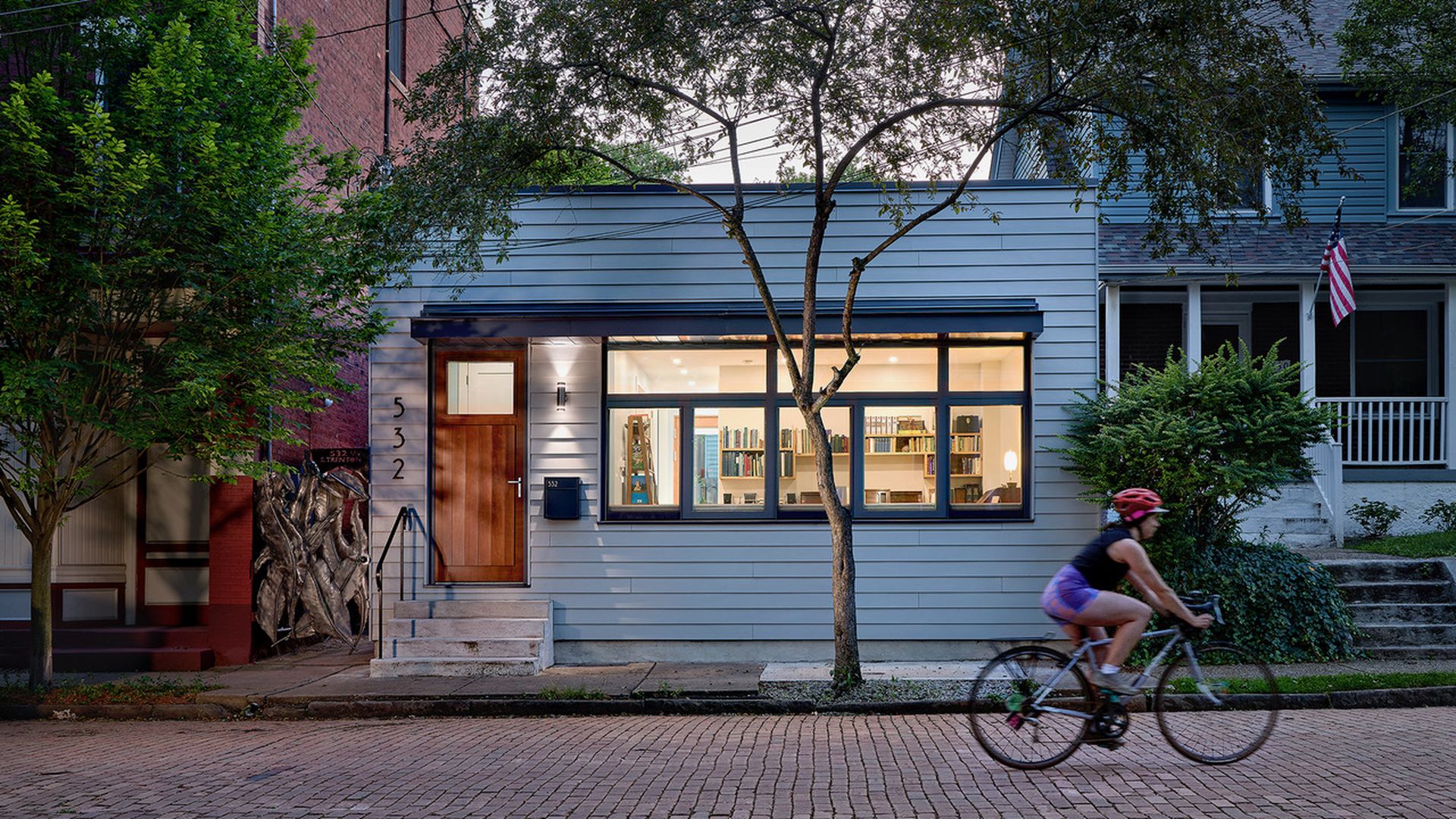 Evening view of a modern, light blue house with large windows revealing a well-lit room filled with bookshelves. A cyclist wearing a red helmet rides past on a brick street.