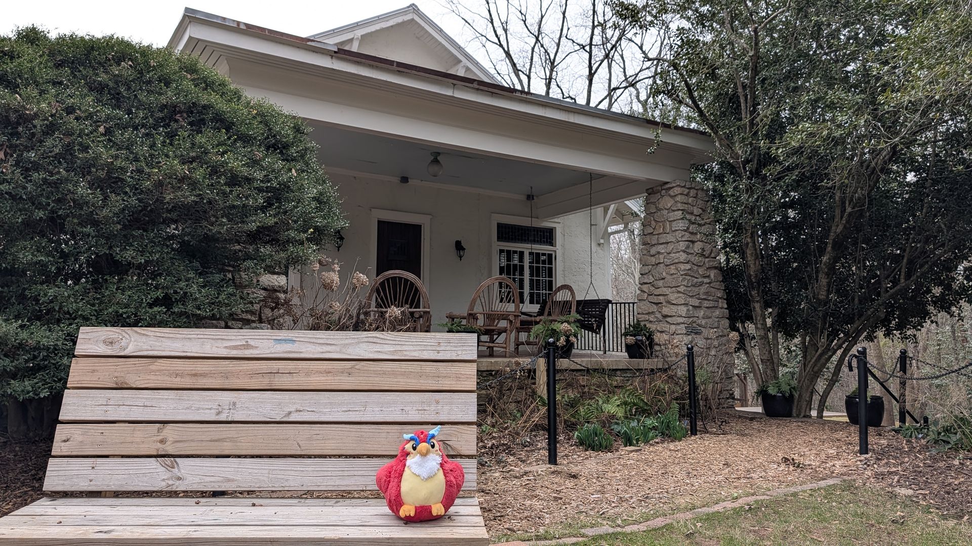 Front porch of a white house with stone columns and a wooden deck. A colorful plush turkey sits on a large wooden step, with shrubs, trees, and fallen leaves around.