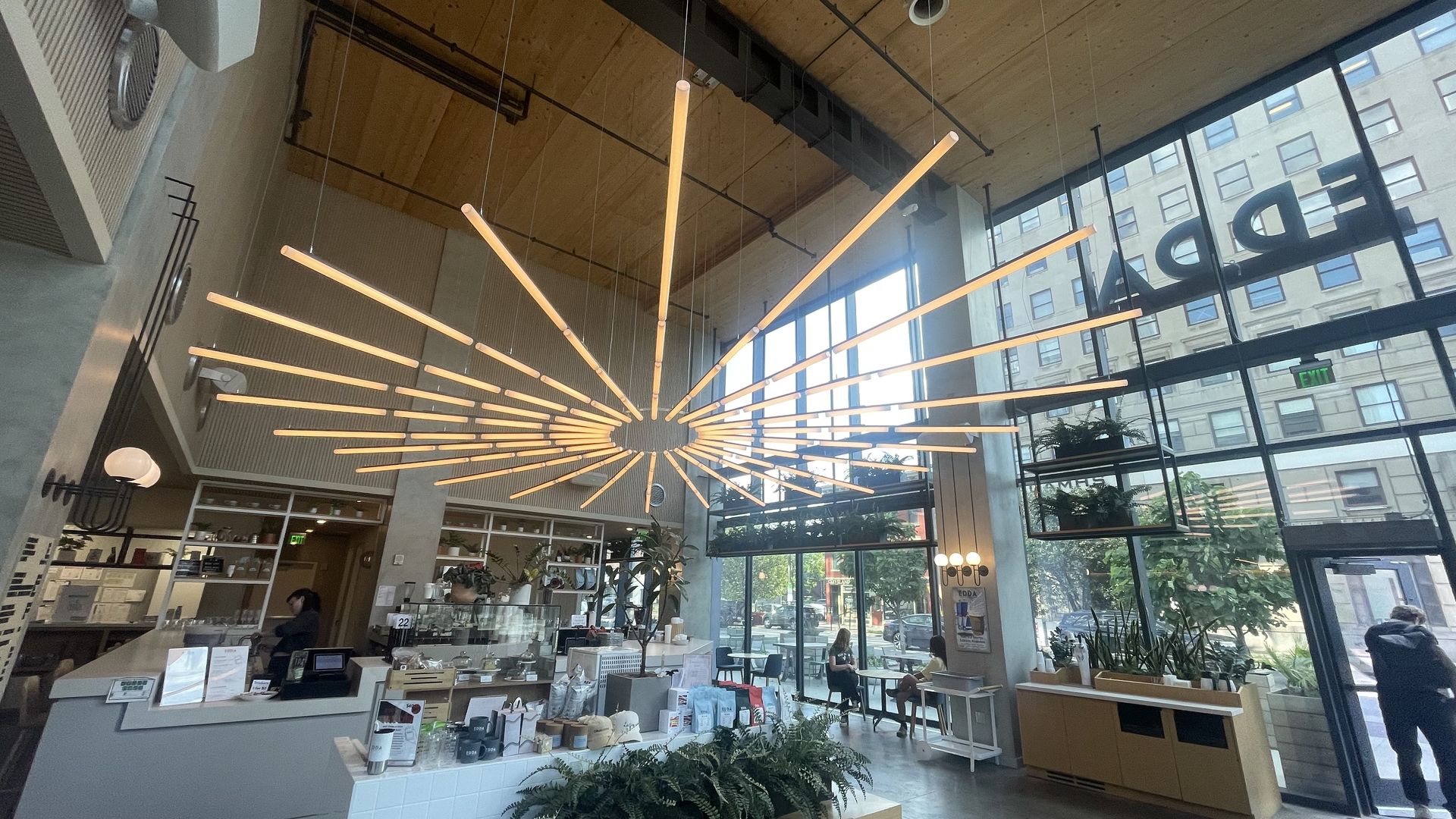 Modern cafe interior with large radial light fixture hanging from wooden ceiling, tall windows, plants, and people sitting at tables. "EDDA" signage visible on the window.
