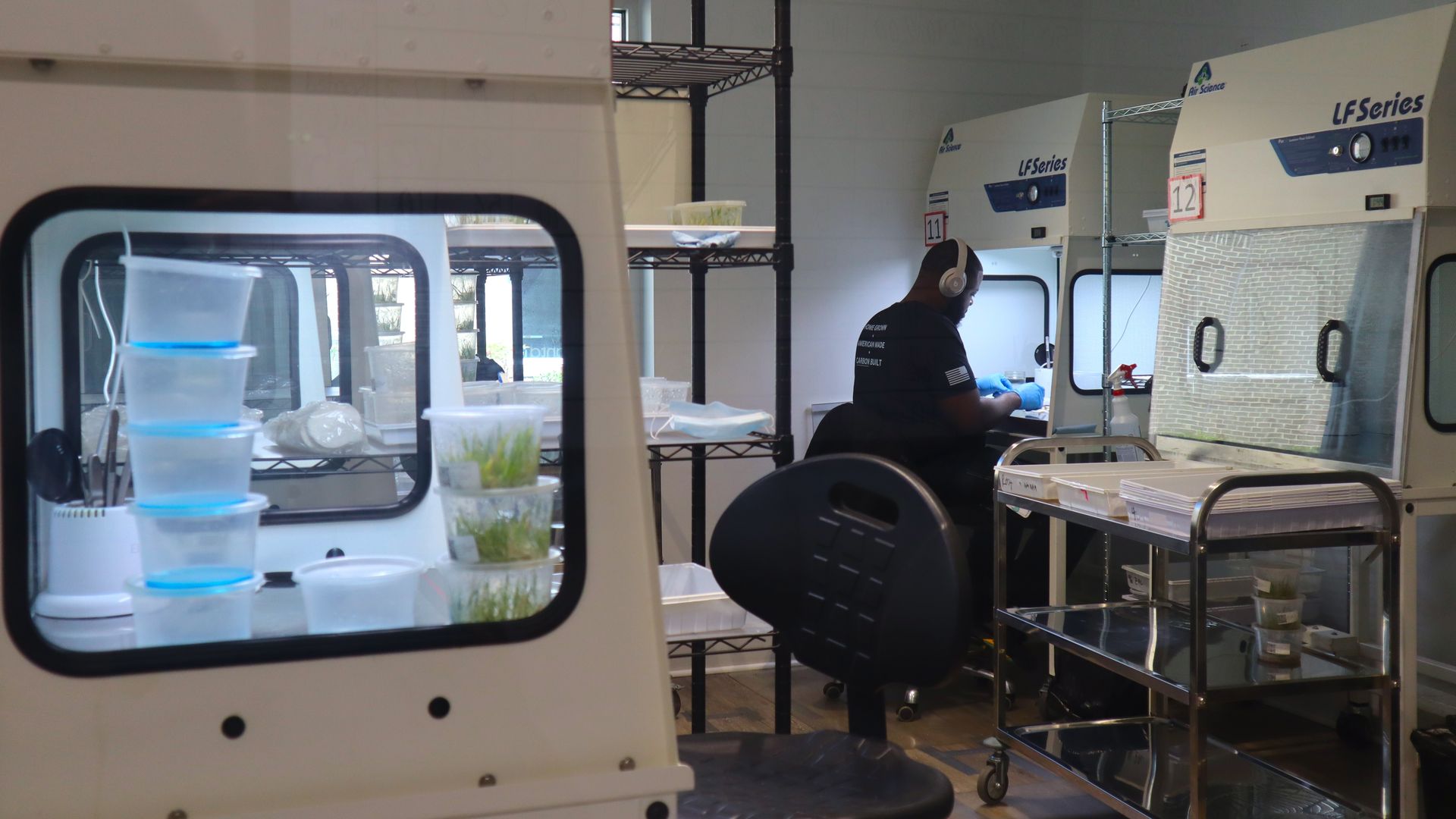 Lab scene with a person wearing headphones and blue gloves at a workstation beside LF Series incubators; foreground shows a white windowed machine with stacked cups of plant tissue and blue liquid.