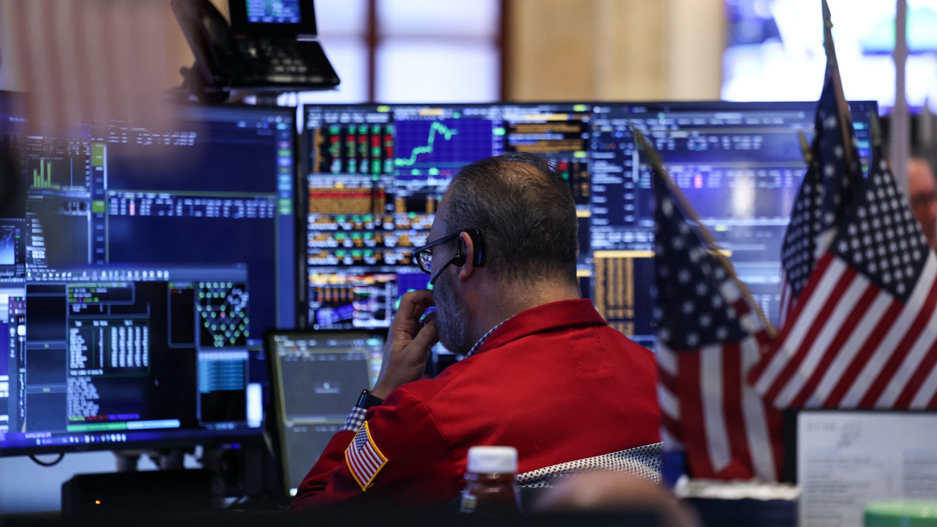 A man in a red jacket and glasses, facing away from the camera and toward a bank of screens showing stock market data. 