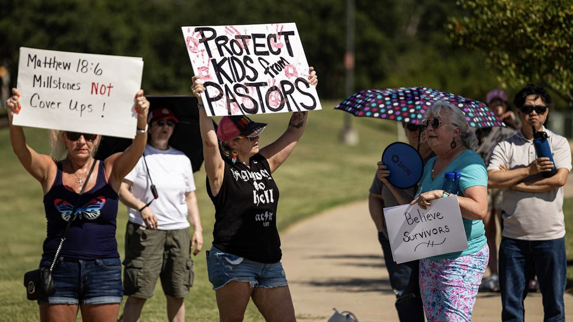 Protesters stand outside holding signs saying "Protect kids from pastors" and "Believe survivors"