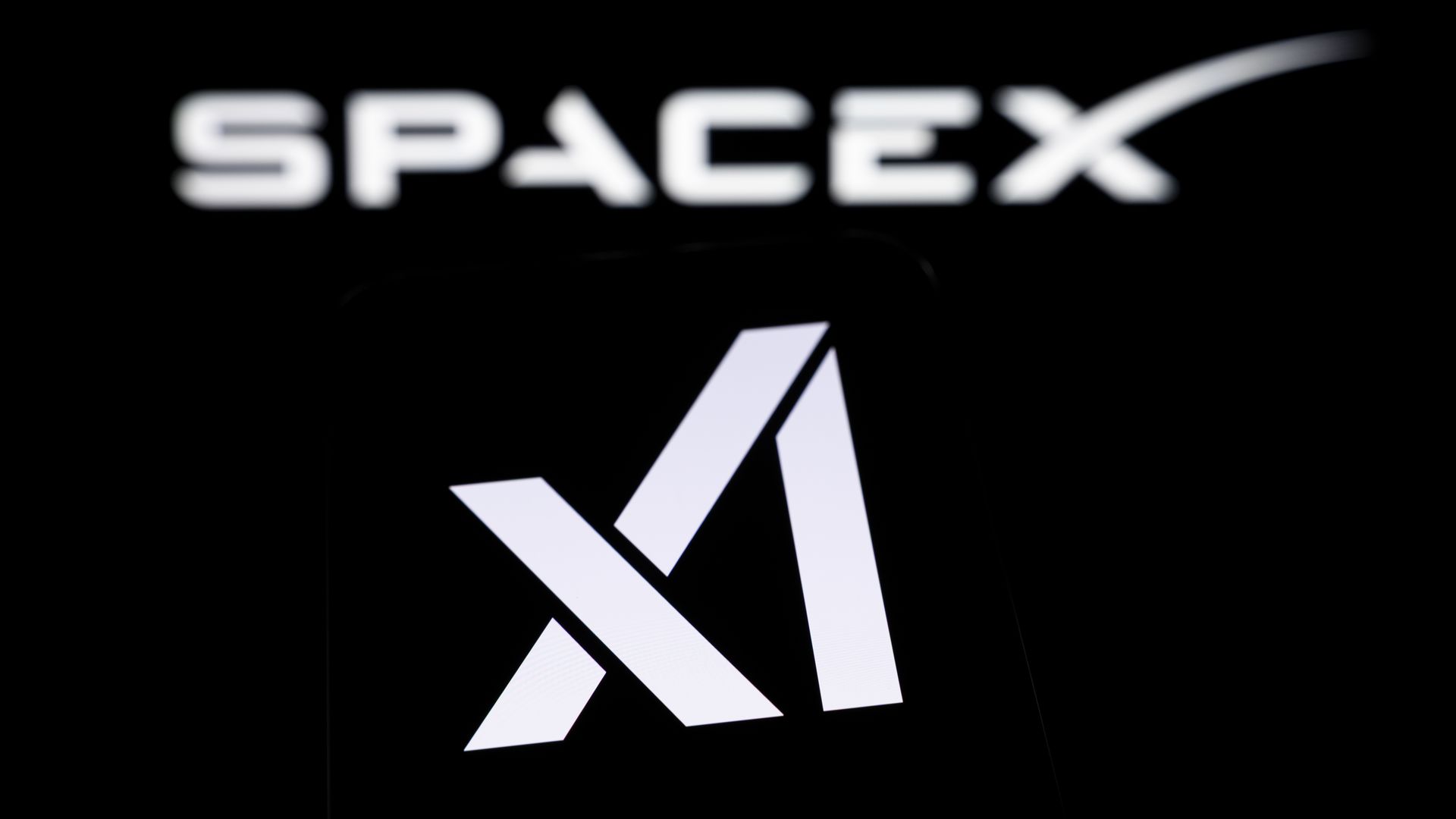 Close-up of a SpaceX logo on a black background: the white word "SPACEX" at the top and a bold, white, diagonal X-shaped symbol below.