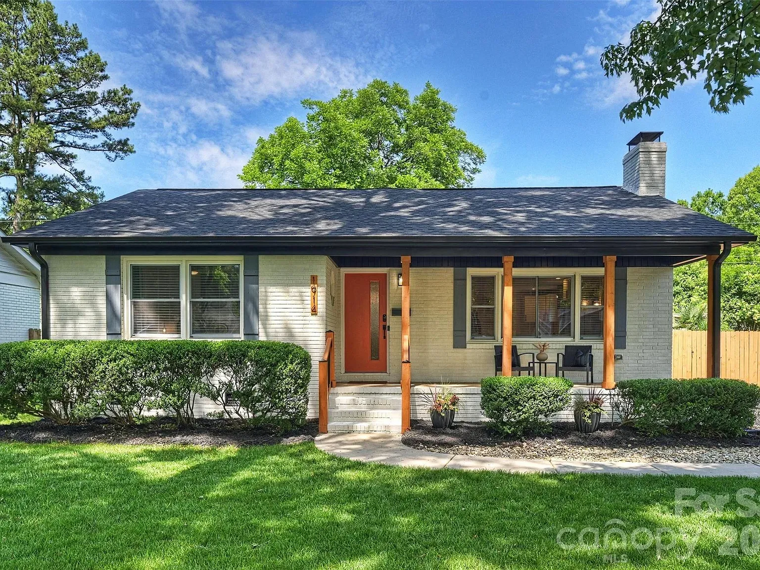 hot homes june 5 2026. Light brick ranch-style house with a dark roof, orange front door, and two wooden porch columns. Front shrubs, a small seating area, and a green lawn under a blue sky.