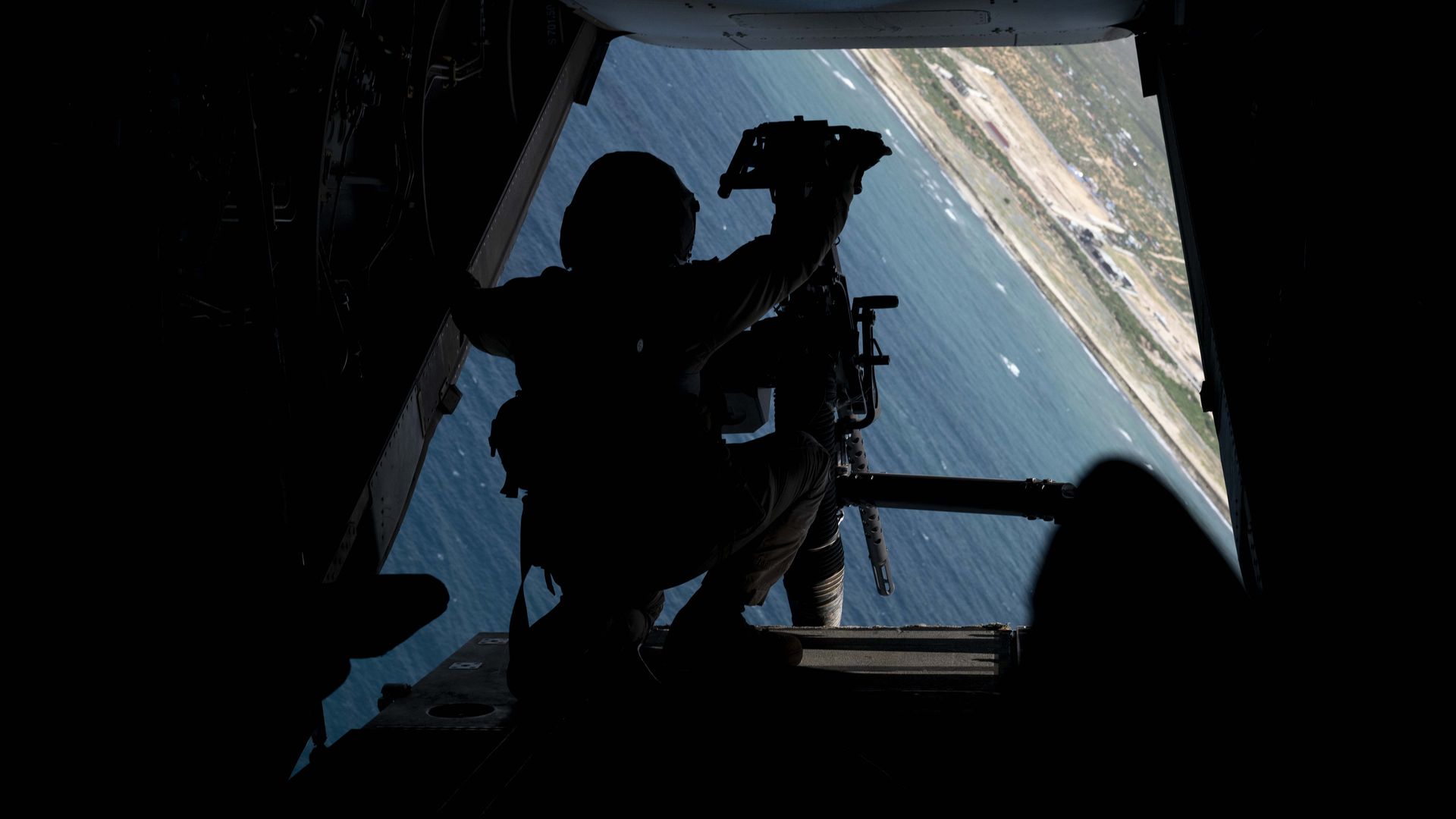 A Marine is crouched behind a gun aboard an airborne Osprey aircraft. Below, waves and Mogadishu, in Africa, are seen.