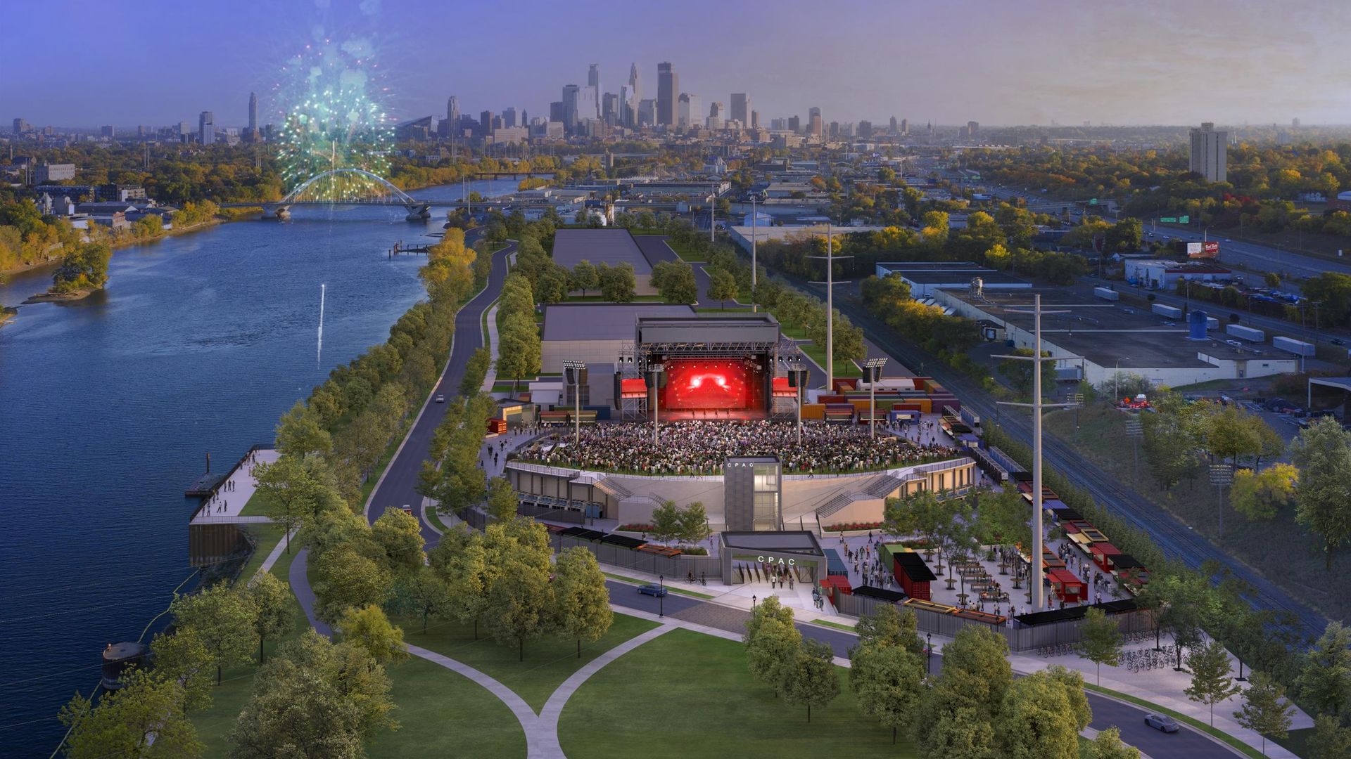 View of a large outdoor concert at CPAC venue near a river, with a stage lit in red, a seated crowd, a green park, and fireworks over a city skyline in the background at dusk.