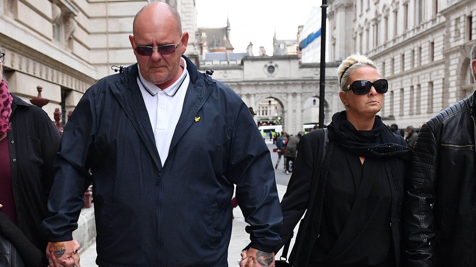 The parents of Harry Dunn dressed in black and holding hands walking down a street.