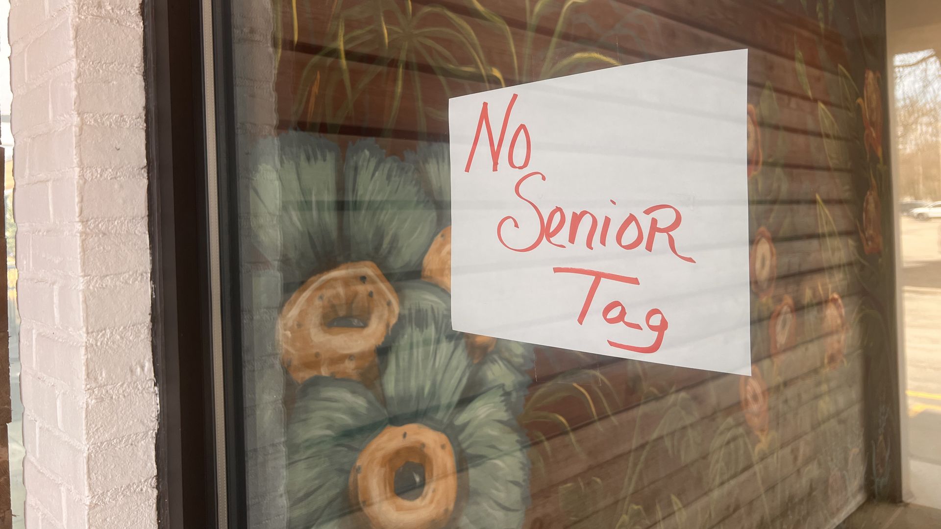 Storefront window with a white sign reading "No Senior Tag" in red cursive; background shows pumpkins and a plant mural with sunlight glare on the glass.