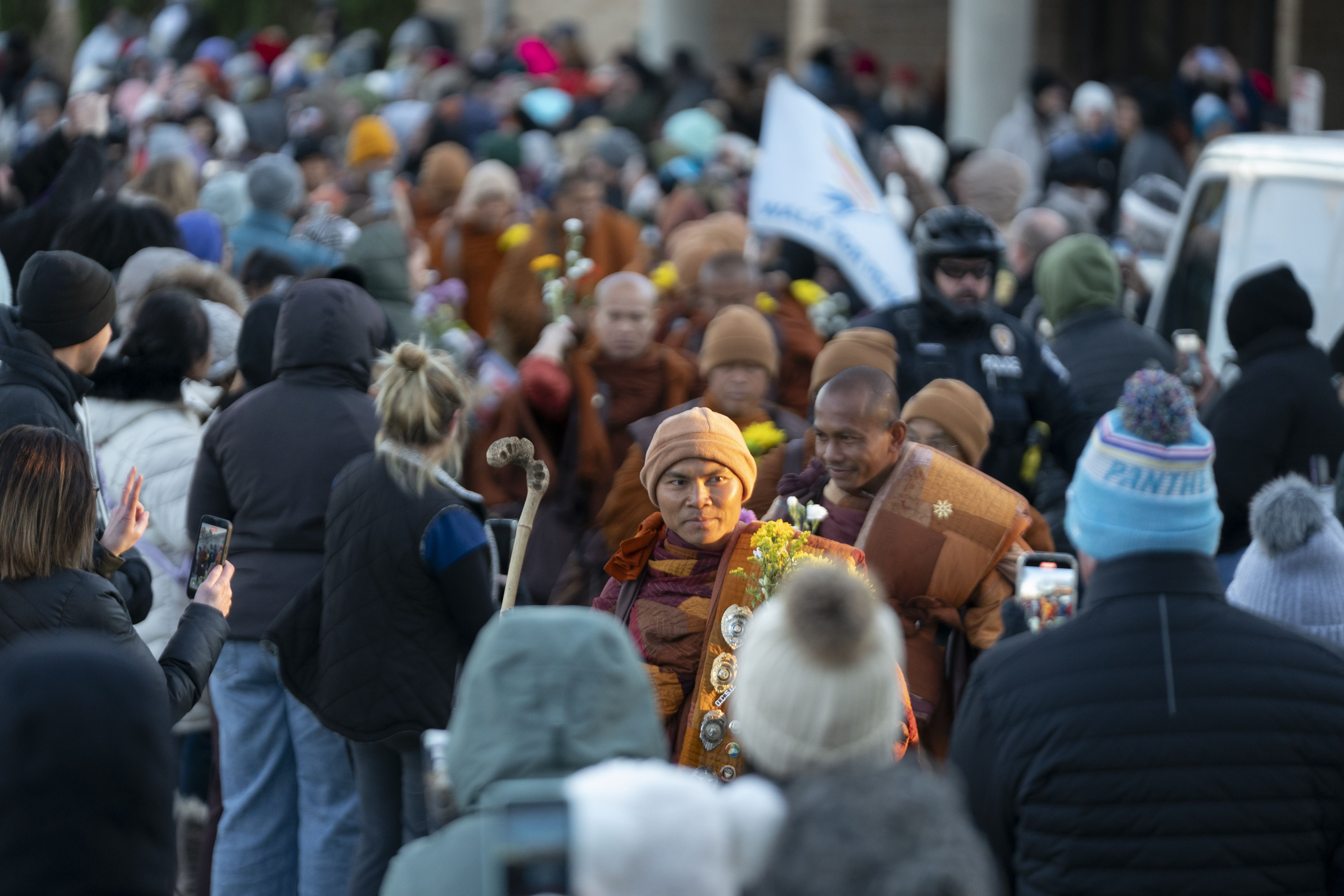 Crowd watching a procession of monks in orange robes, some holding flowers. People in winter clothes take photos. Police officer stands nearby. One monk in focus wears badges on his robe.