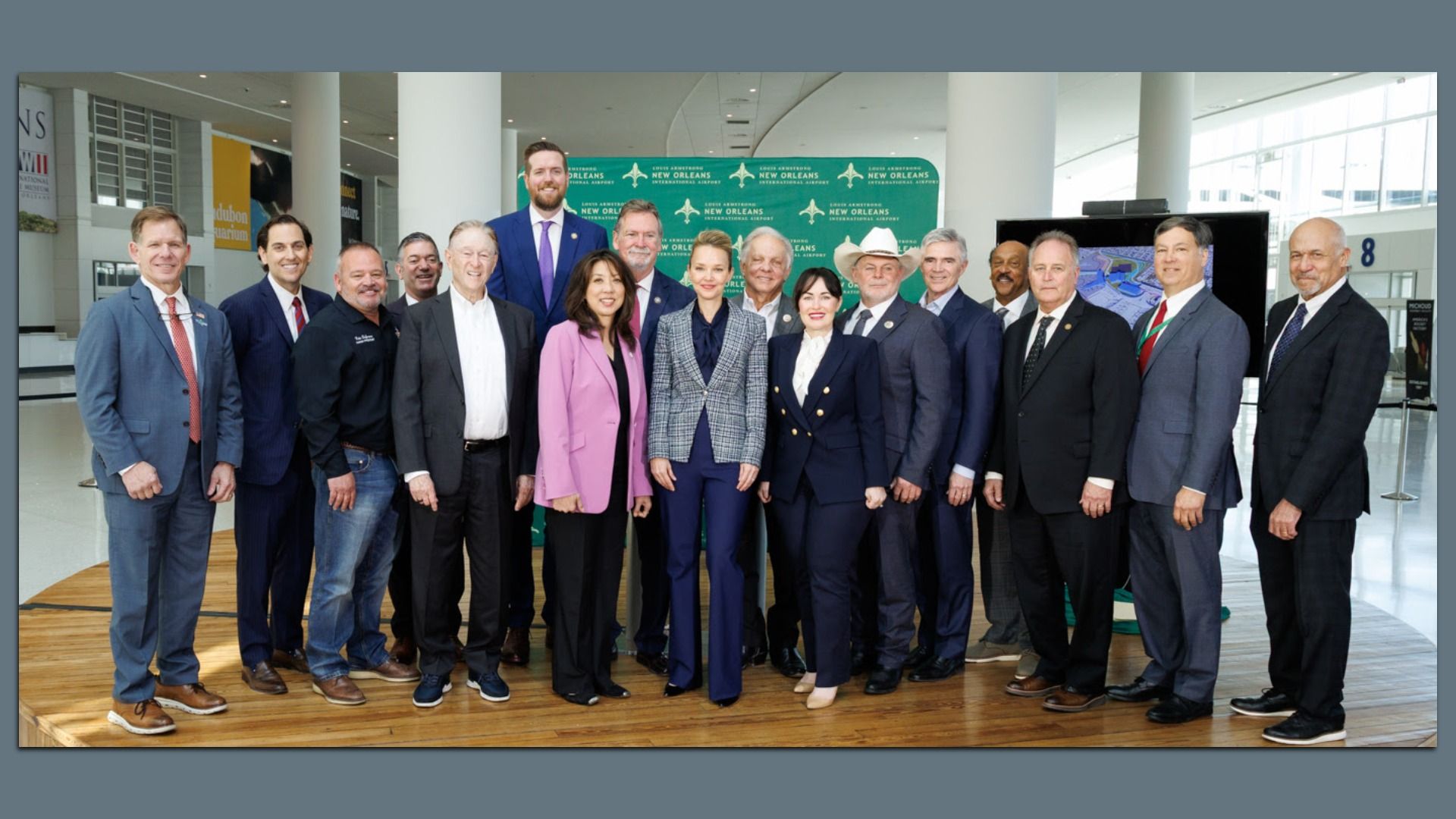 Group of about 20 professionals in business attire posing for a photo on a wooden floor, in front of a green backdrop with New Orleans logos at an airport terminal.