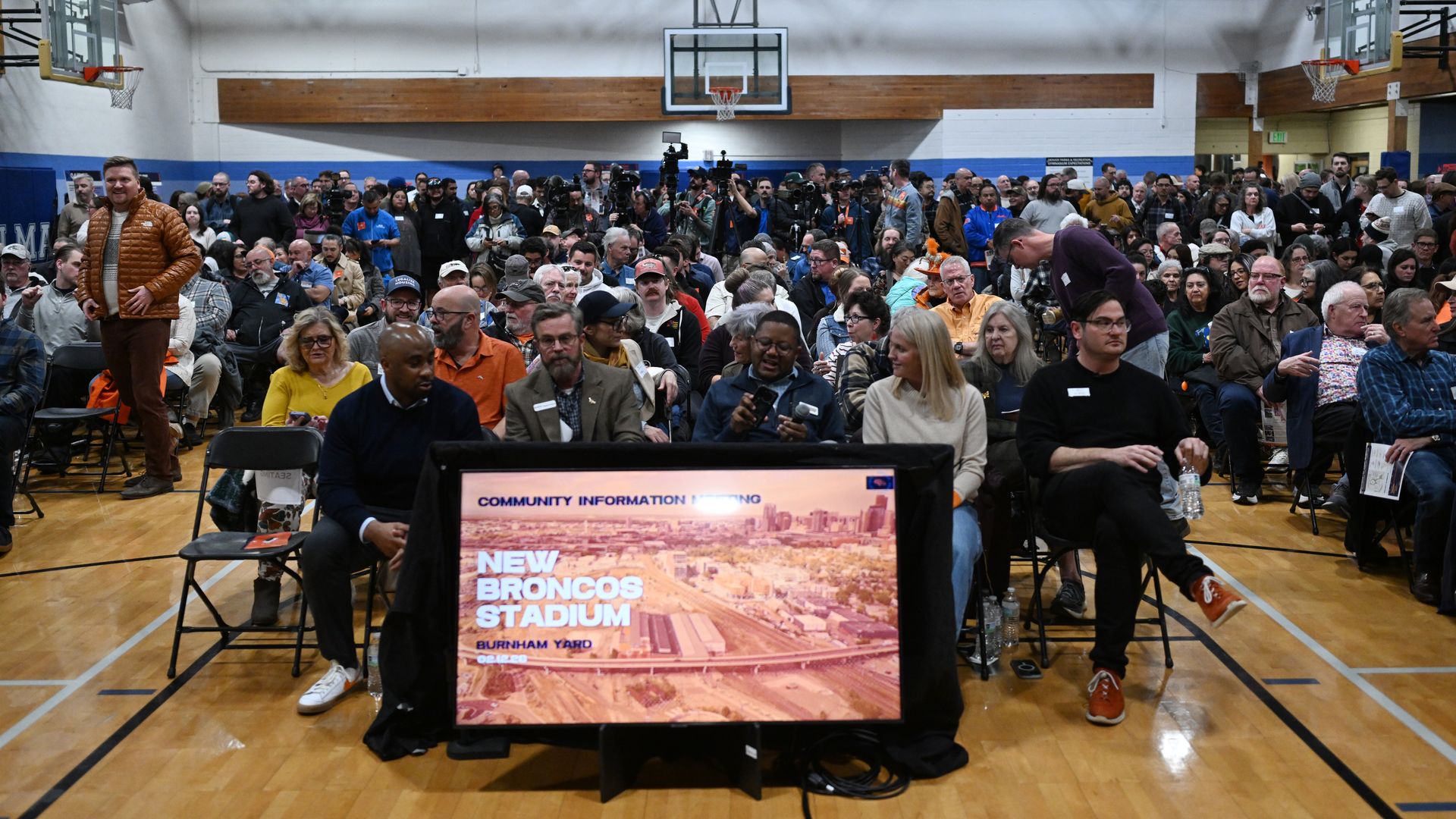 Busy gymnasium full of a large crowd and media, with cameras in the center. A screen shows New Broncos Stadium, flanked by basketball hoops and a wood-beamed ceiling.
