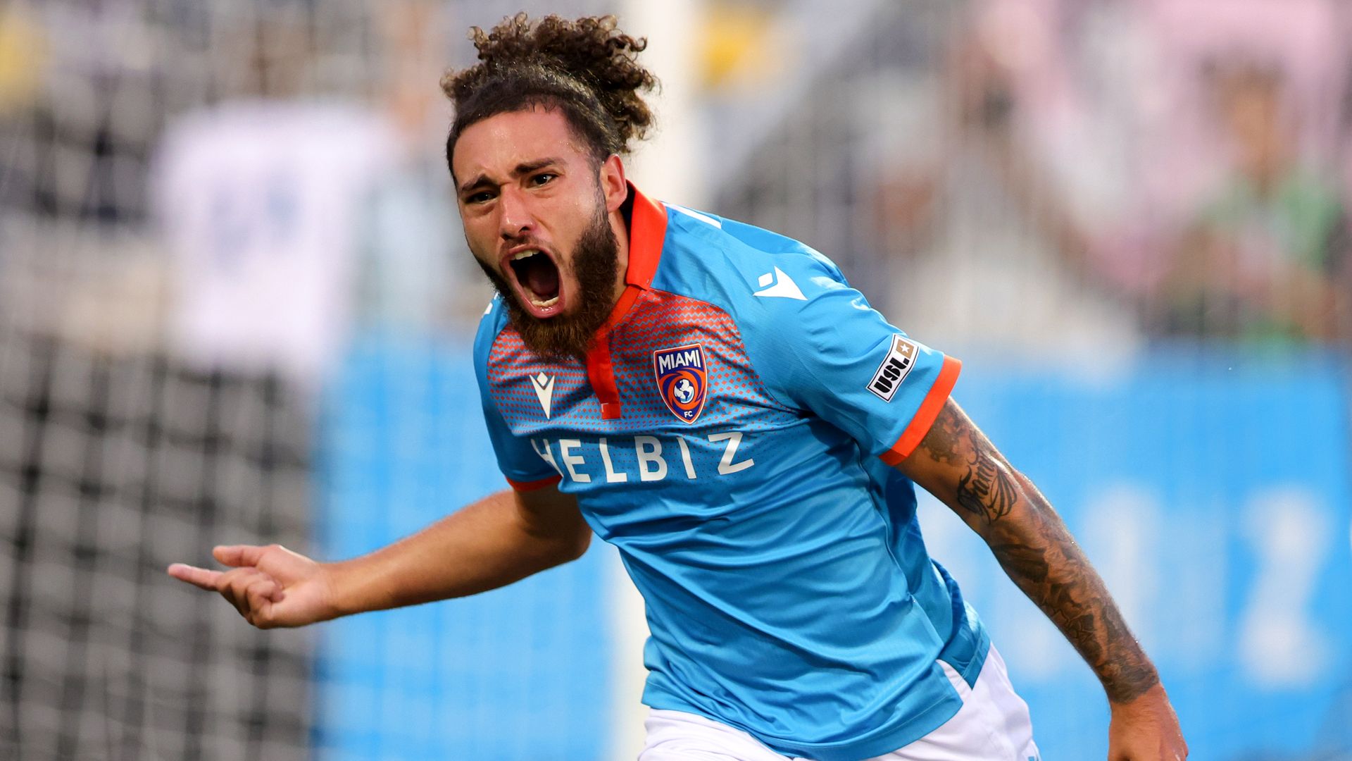 A soccer player with curly hair pulled into a ponytail shouts while playing a game.