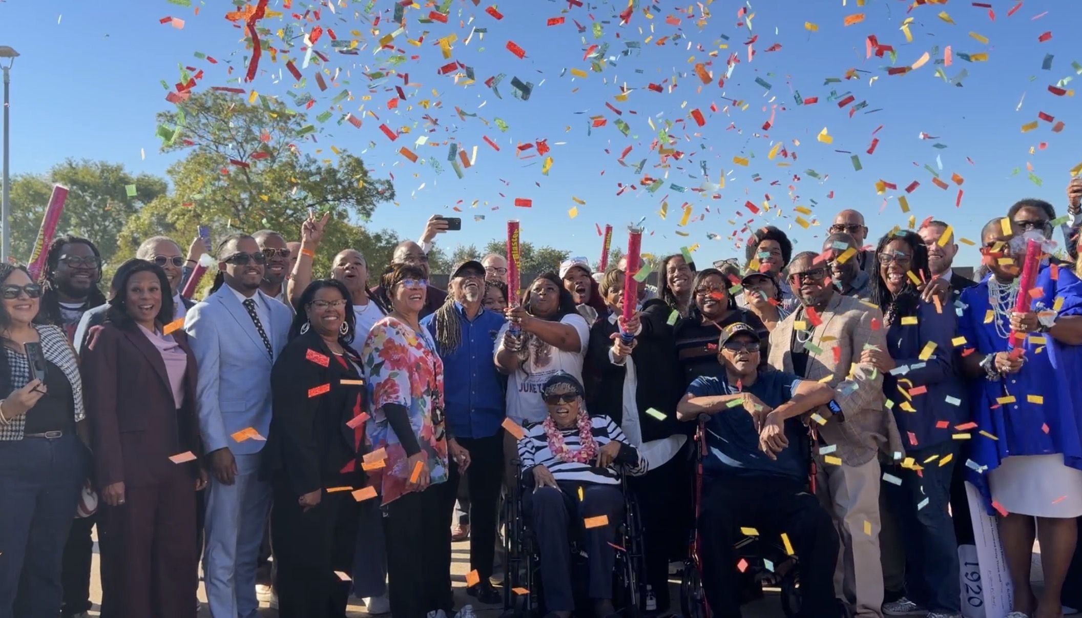 Large, diverse group celebrating outdoors, smiling as colorful confetti showers down from party poppers over a clear blue sky.