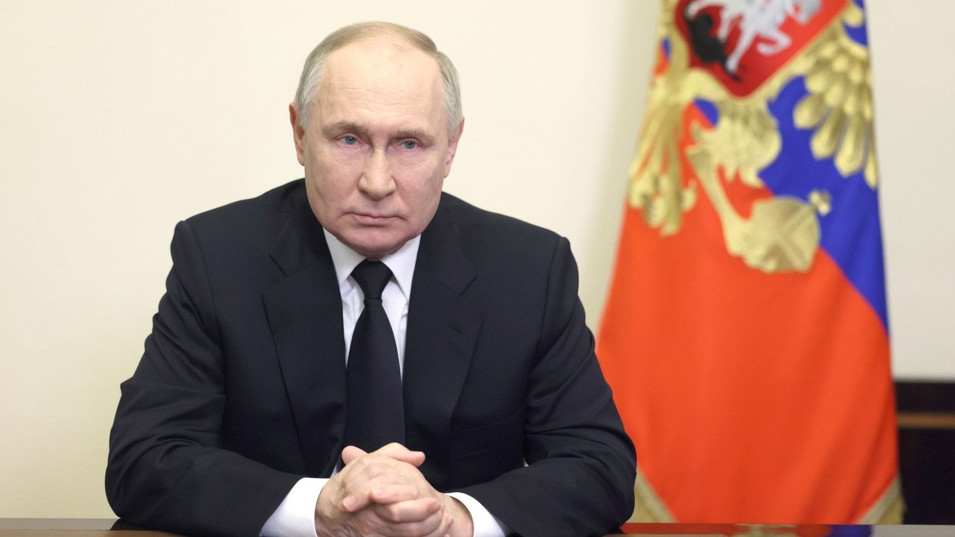 Russian President Vladimir Putin sits with hands clasped and a flag behind him. 