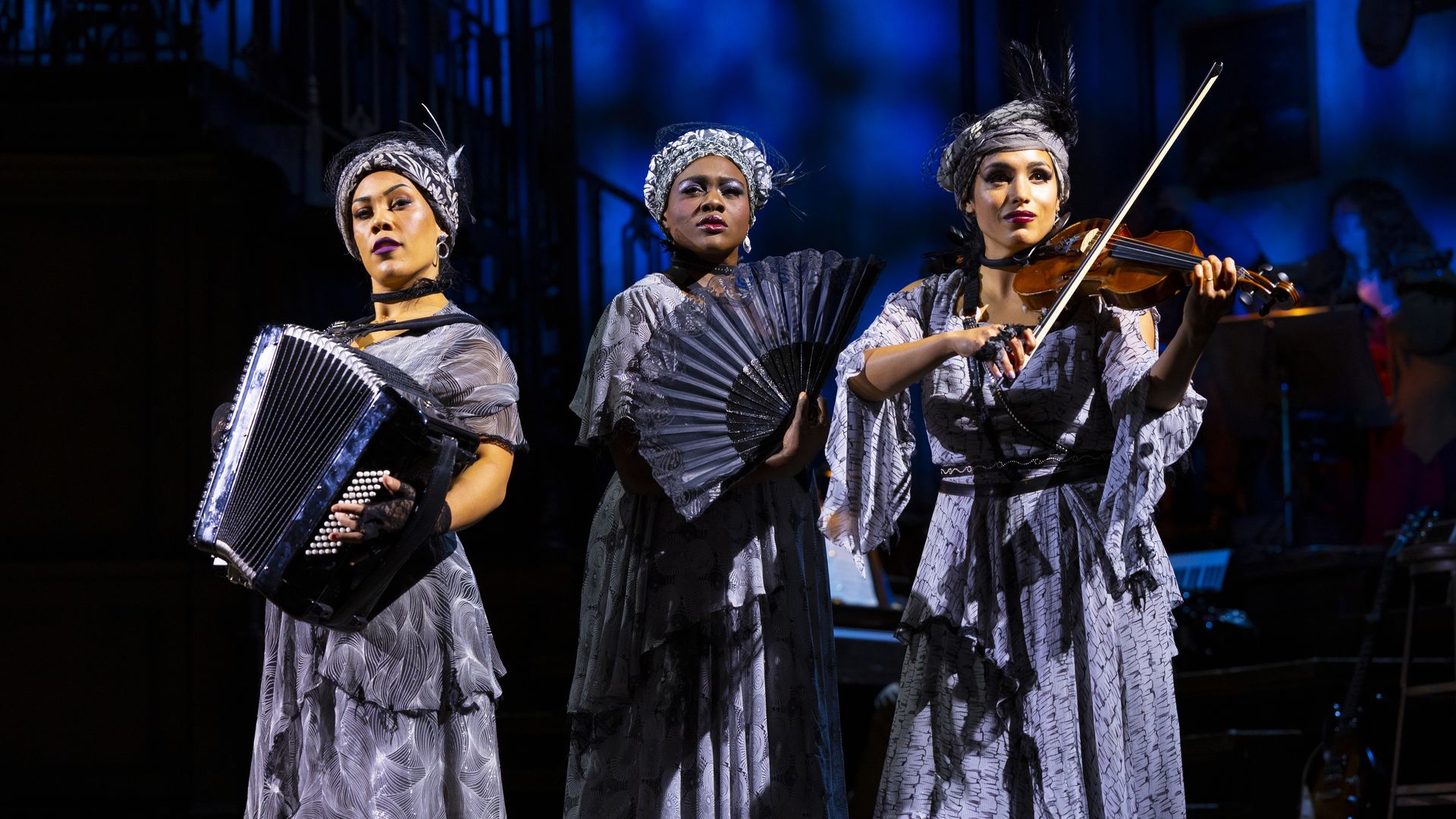 Three actresses in purple dresses play instruments and pose on stage in the musical Hadestown.