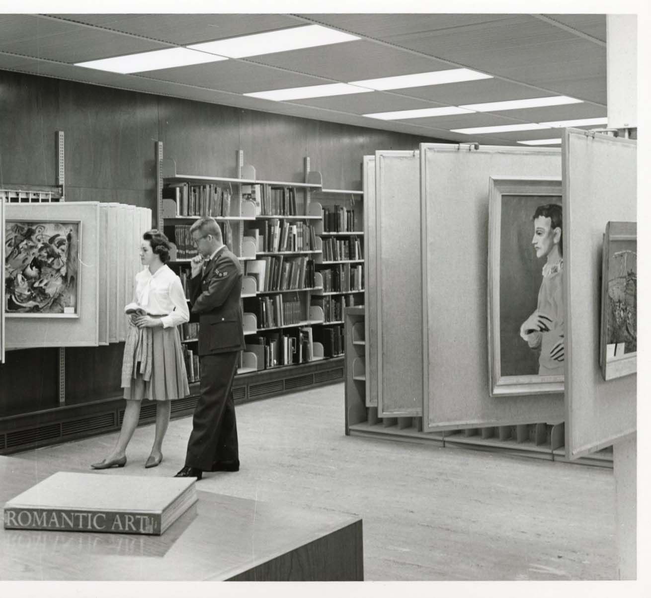 Black-and-white photo of a man in a military uniform and a woman in a white blouse and skirt examining a painting in a gallery-like library. Books line the back; large art panels stand nearby; pedestal reads "ROMANTIC ART".